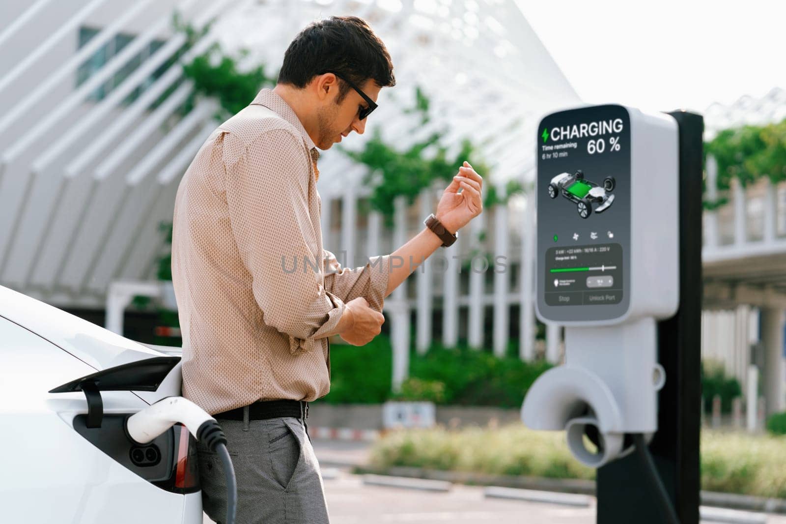 Young man put EV charger to recharge electric car's battery from charging station in city commercial parking lot. Rechargeable EV car for sustainable environmental friendly urban travel. Expedient