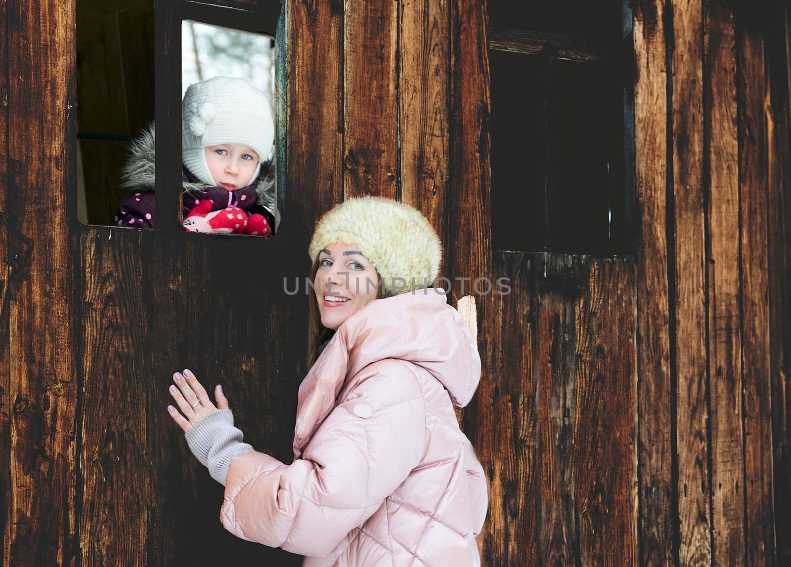 Pretty mother with a child in the window of a wooden toy house in a winter park by jovani68