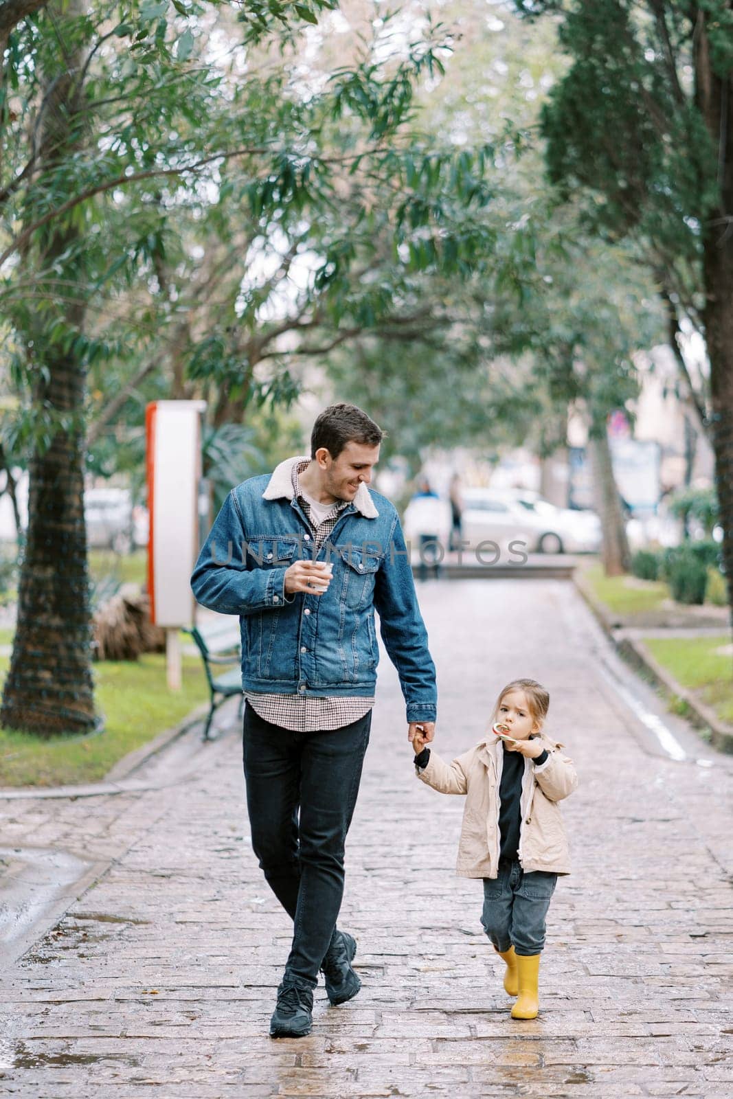Dad with a glass of coffee walks down the street holding the hand of a little girl with a candy cane. High quality photo