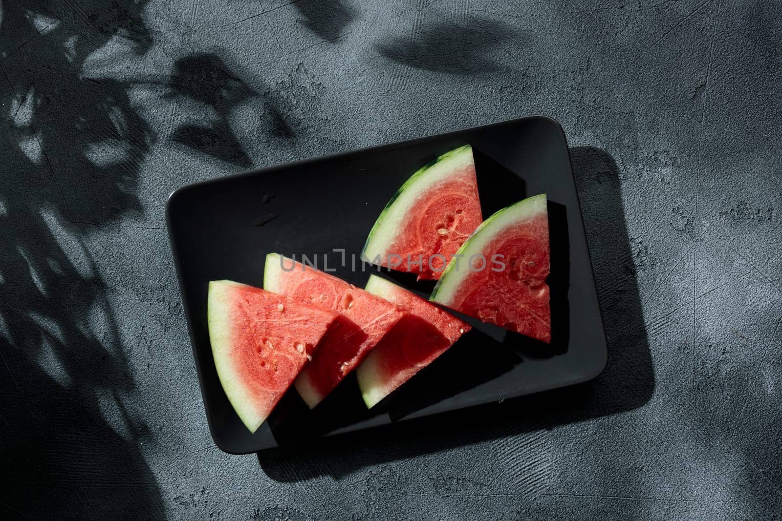 Slices of juicy watermelon on the black plate on the concrete background with summer sun shadows. Copy space. Top view.