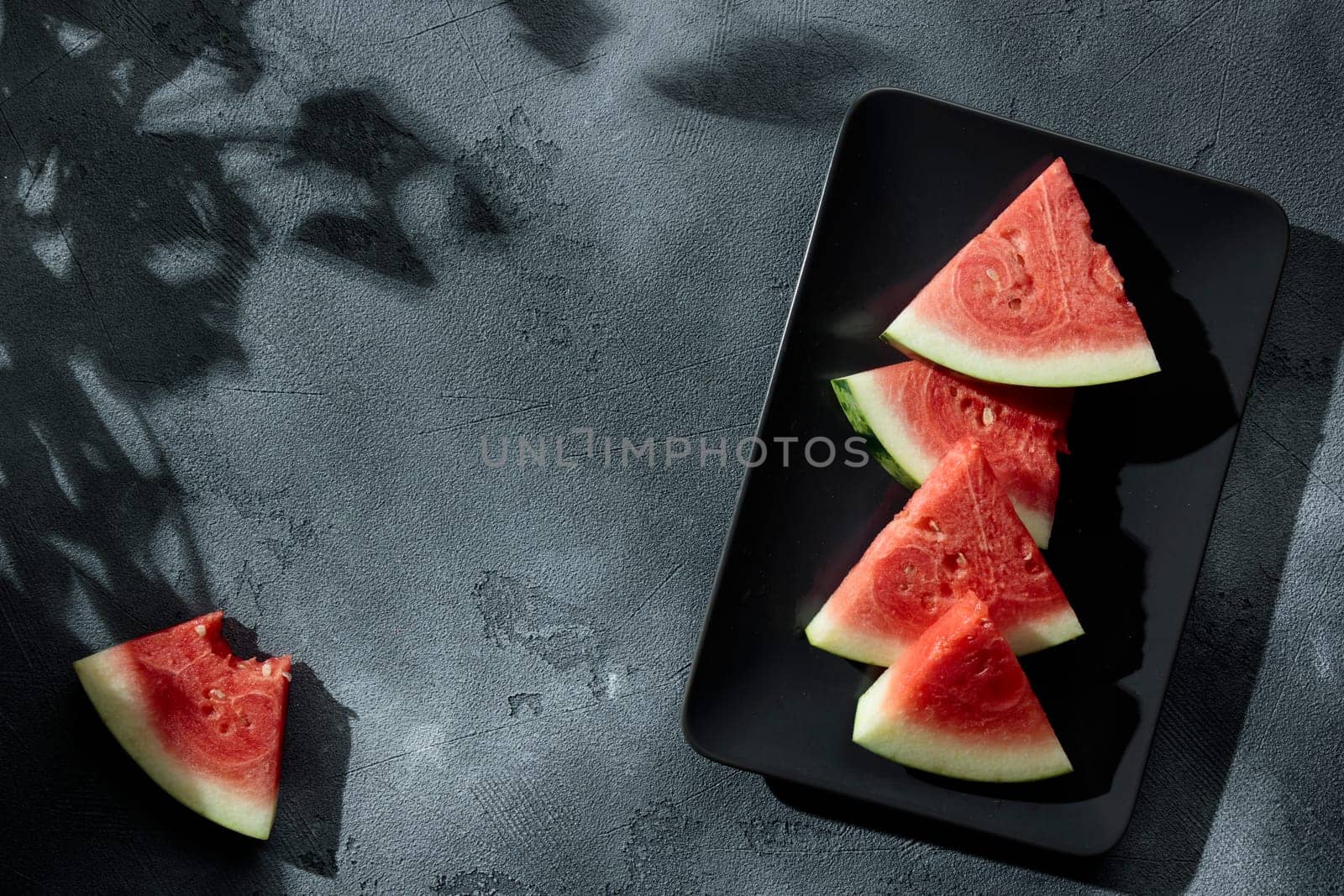 Slices of juicy watermelon on the black plate on the concrete background with summer sun shadows. Copy space. Top view.