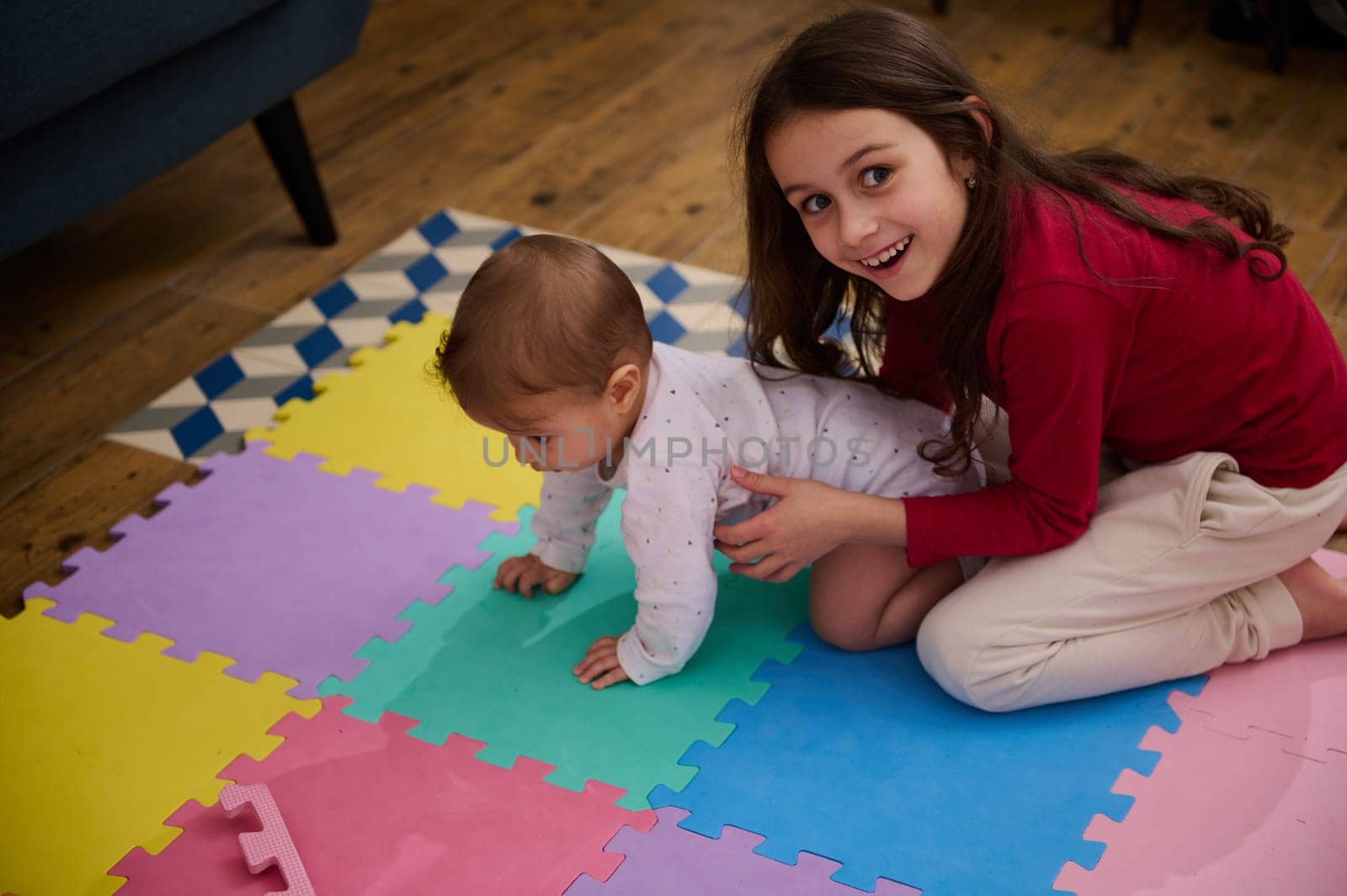 Adorable little girl smiling looking at camera, expressing positive emotions and happiness helping her brother, a cute baby boy on crawling on colorful puzzle carpet at home. Children. Growth. Family by artgf
