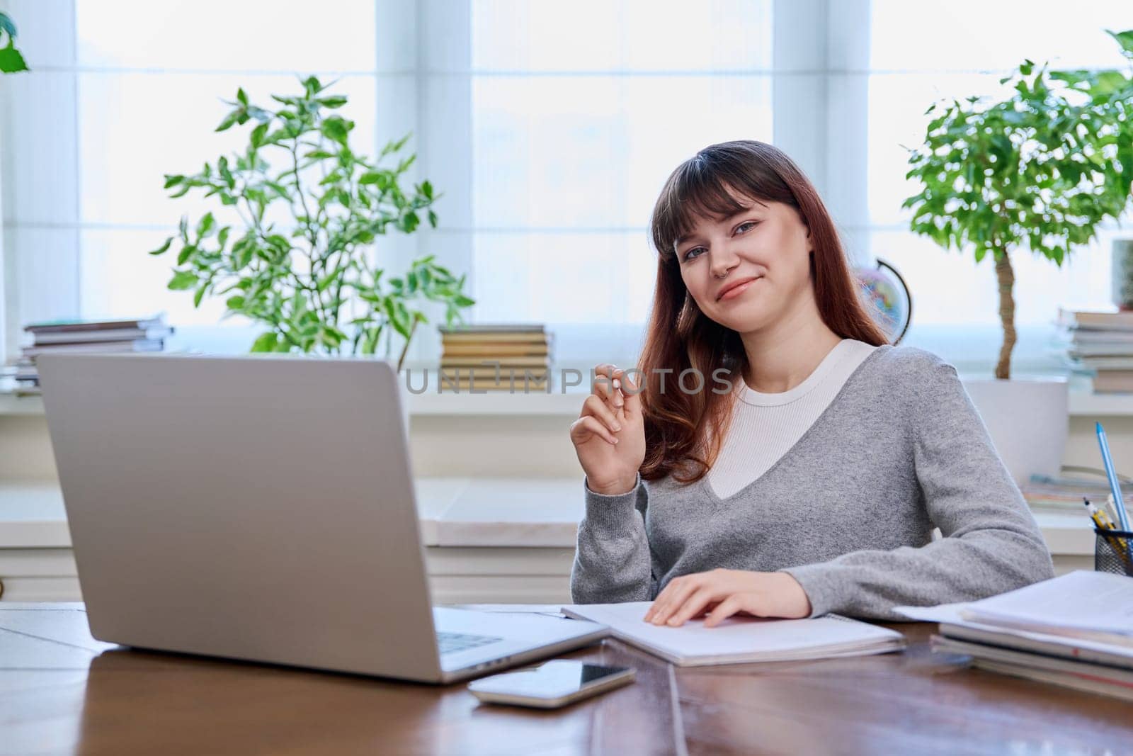 Young female college student studying at home at desk using computer laptop, writing in notebook, smiling looking at camera. E-learning, education, technology, knowledge, youth concept