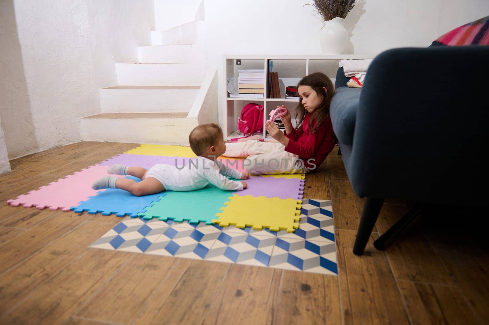 Side portrait of a cute little child girl, loving caring sister playing with her baby boy brother on a colorful puzzle carpet at cozy home interior. Kids. Happy family and World Children's Day concept by artgf