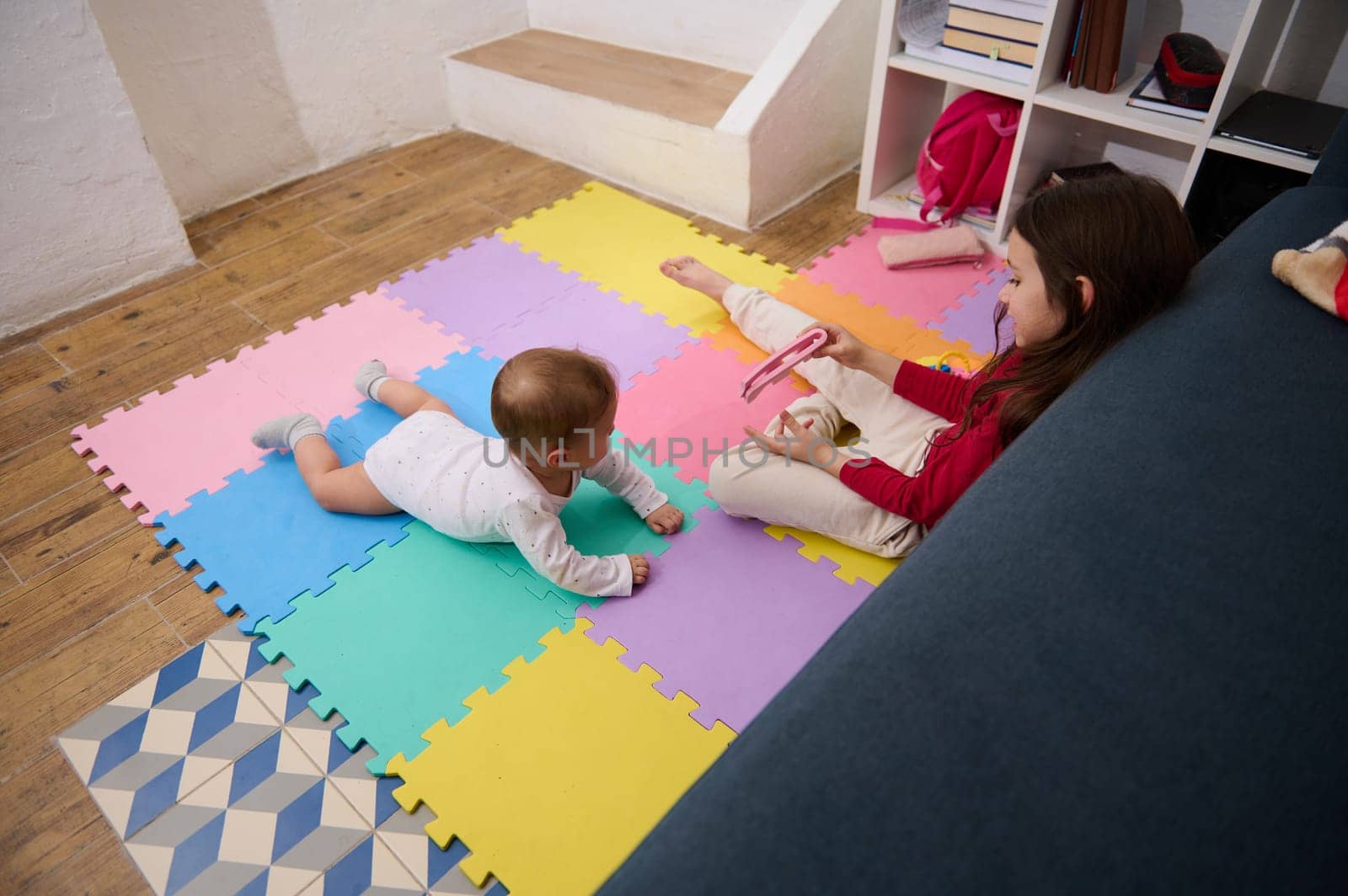 Overhead view of a little child girl, loving caring sister playing with her baby boy brother on a colorful puzzle carpet at cozy home interior. Kids. Happy family and World Children's Day concept by artgf