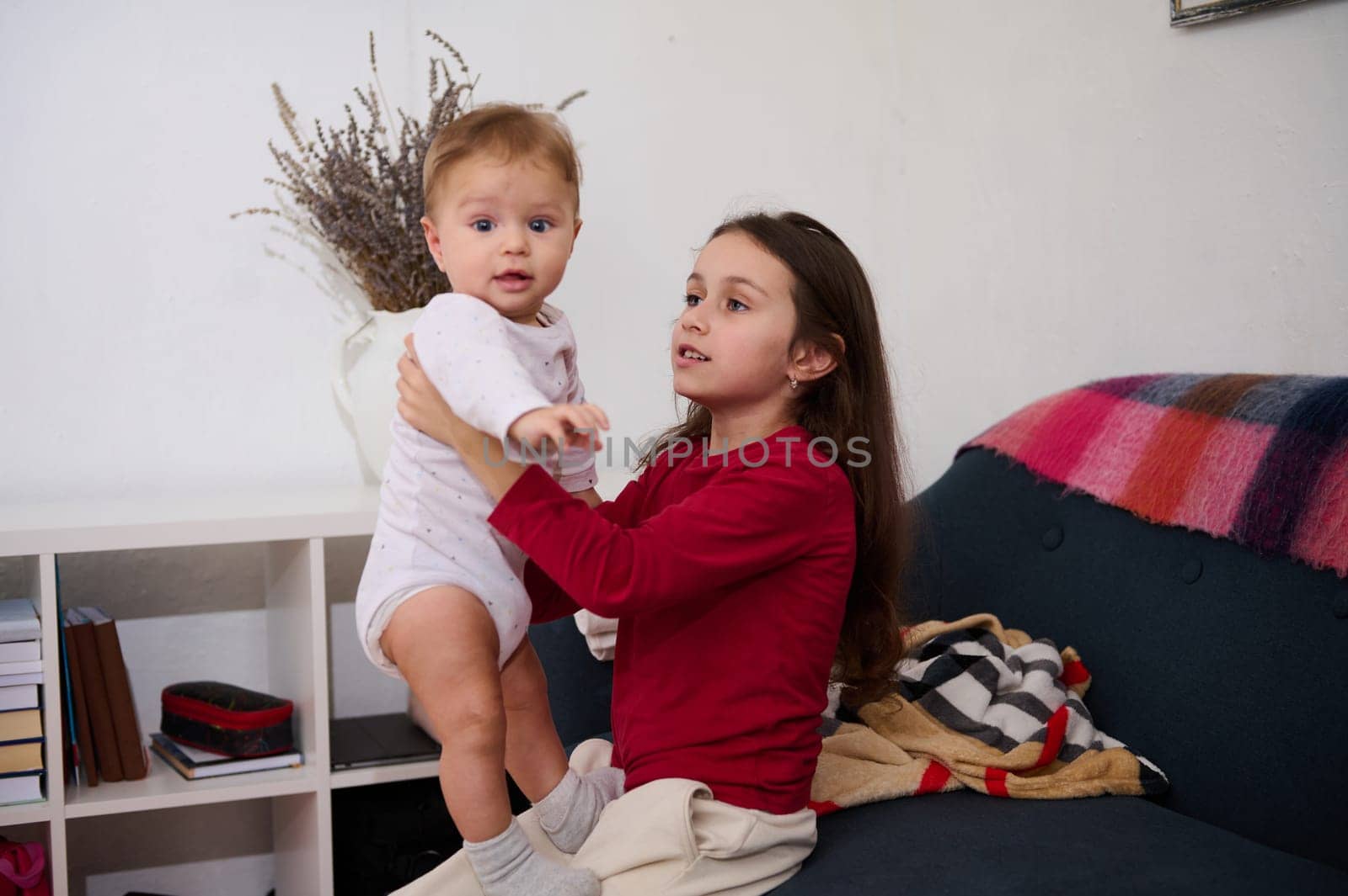Sister holding her brother, an adorable baby boy on white bodysuit, playing together at home. Family relationships and love concept. Adorable little kid girl 6 years old helps her mom with a baby boy