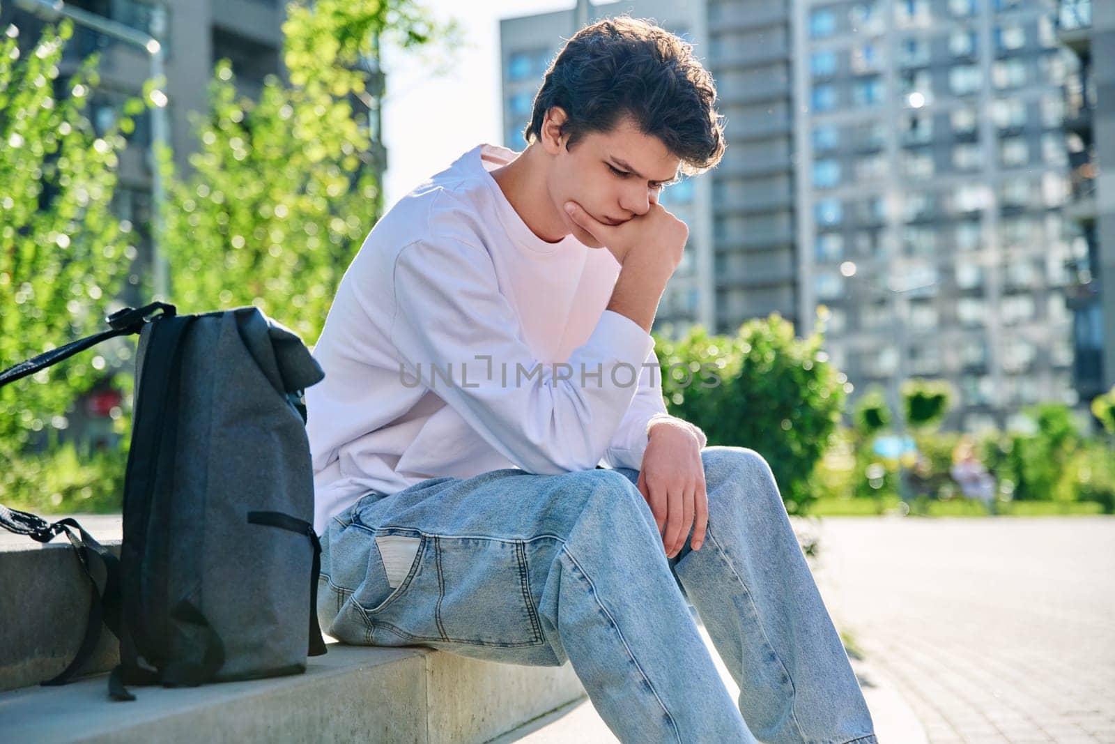 Upset sad unhappy young male sitting outdoor on steps. Guy university college student with backpack sitting, holding head with hands. Problems, difficulties, depression, mental health of young people
