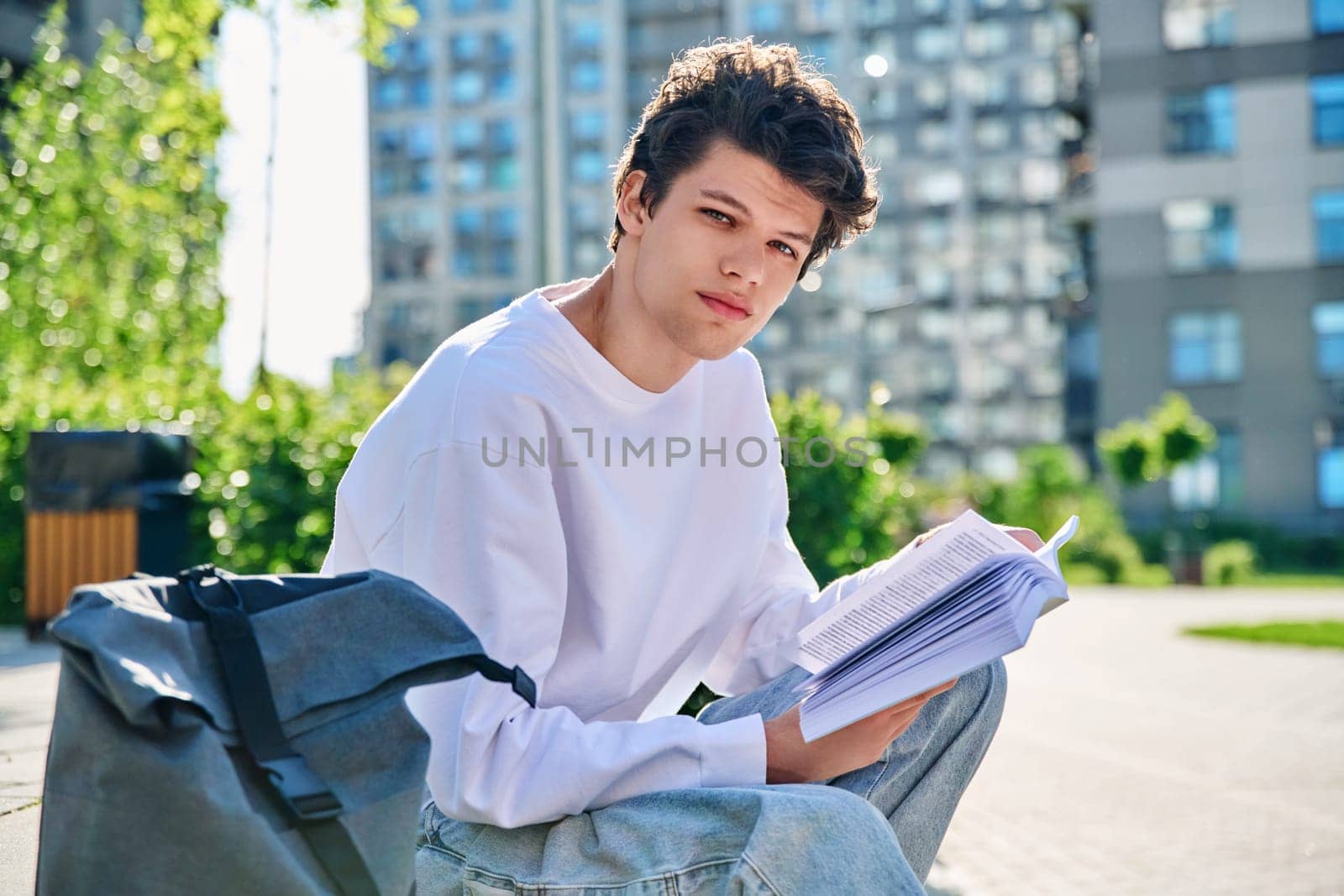Handsome young male university college student with book looking at camera sitting on outdoor steps. Education, knowledge, youth 19-20 years old, literary hobby and leisure