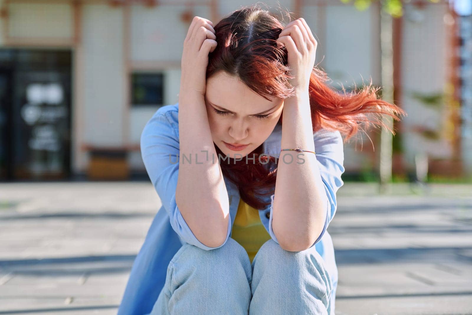 Upset sad unhappy young female sitting outdoor on steps. Girl university college student sitting, holding head with hands. Problems, difficulties, depression, mental health of young people