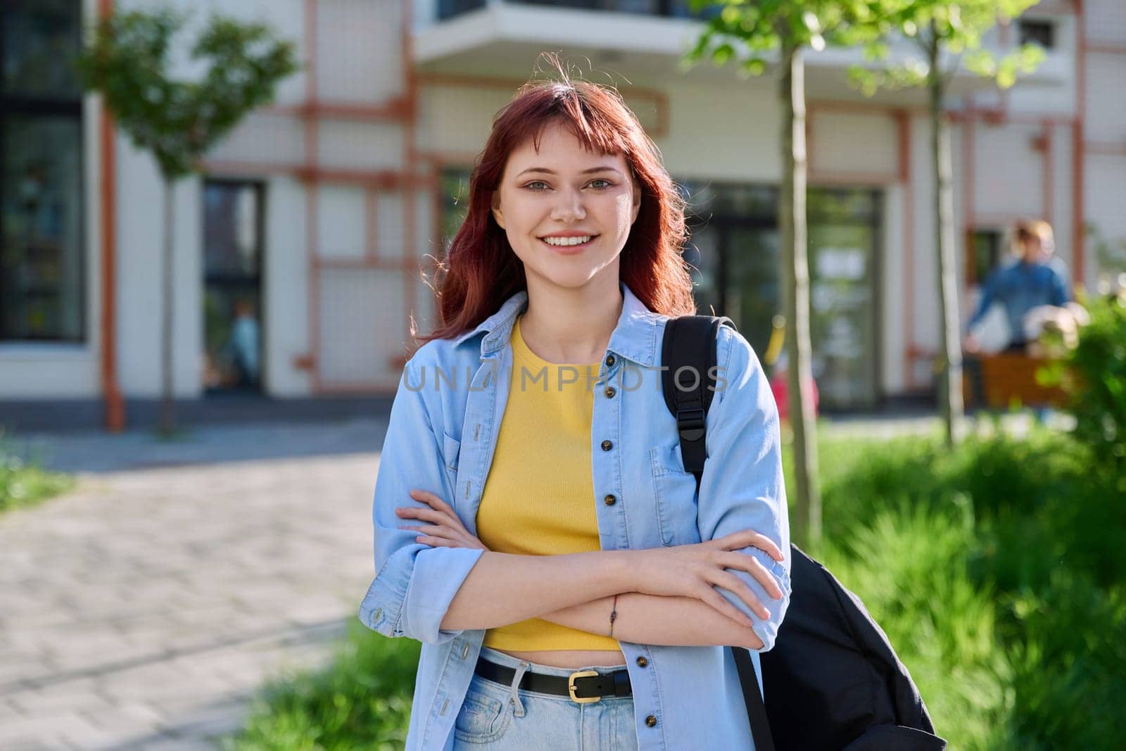 Portrait of young beautiful female college student outdoor, smiling red-haired confident girl with crossed arms backpack looking at camera, educational building background. Education training youth