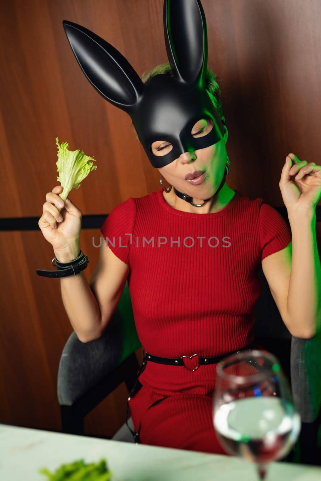 A beautiful girl in a bdsm rabbit mask and a bright red dress eats lettuce leaves promoting a healthy lifestyle and vegetarianism