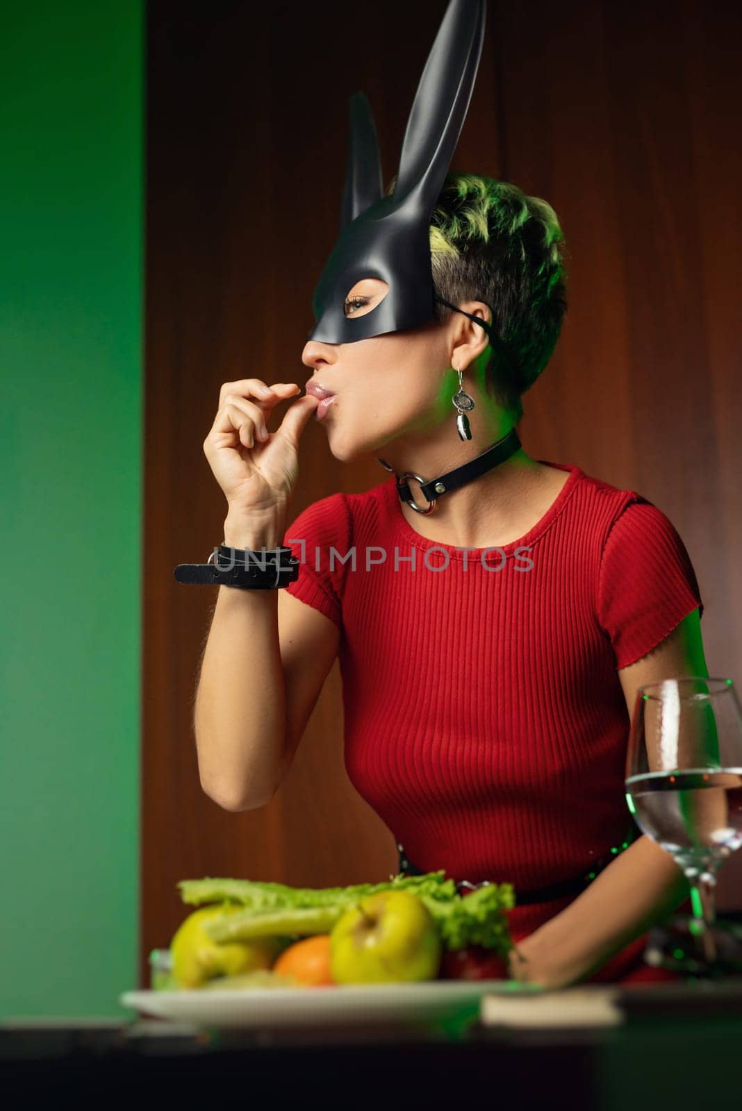 A beautiful girl in a bdsm rabbit mask and a bright red dress eats lettuce leaves promoting a healthy lifestyle and vegetarianism by Rotozey