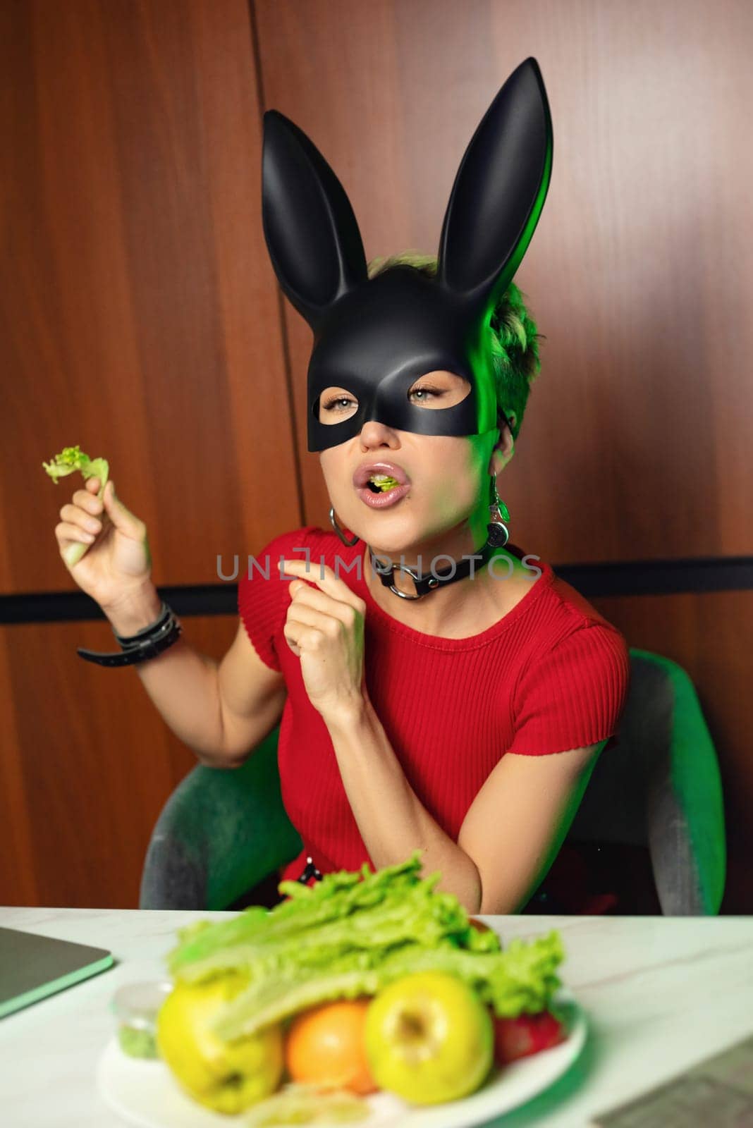 A beautiful girl in a bdsm rabbit mask and a bright red dress eats lettuce leaves promoting a healthy lifestyle and vegetarianism