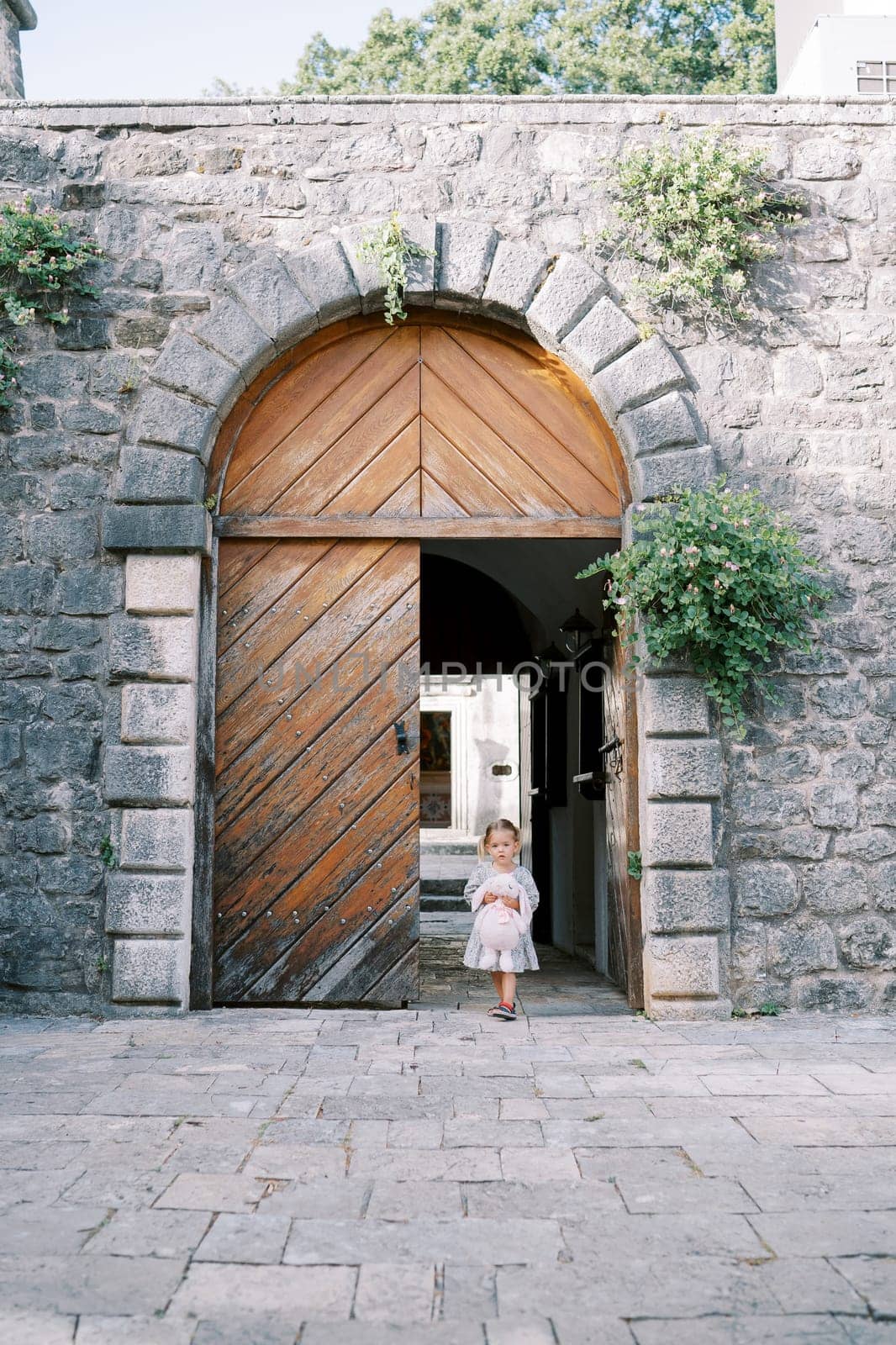 Little girl with a pink rabbit comes out of the arched wooden doors into the courtyard of an ancient castle. High quality photo