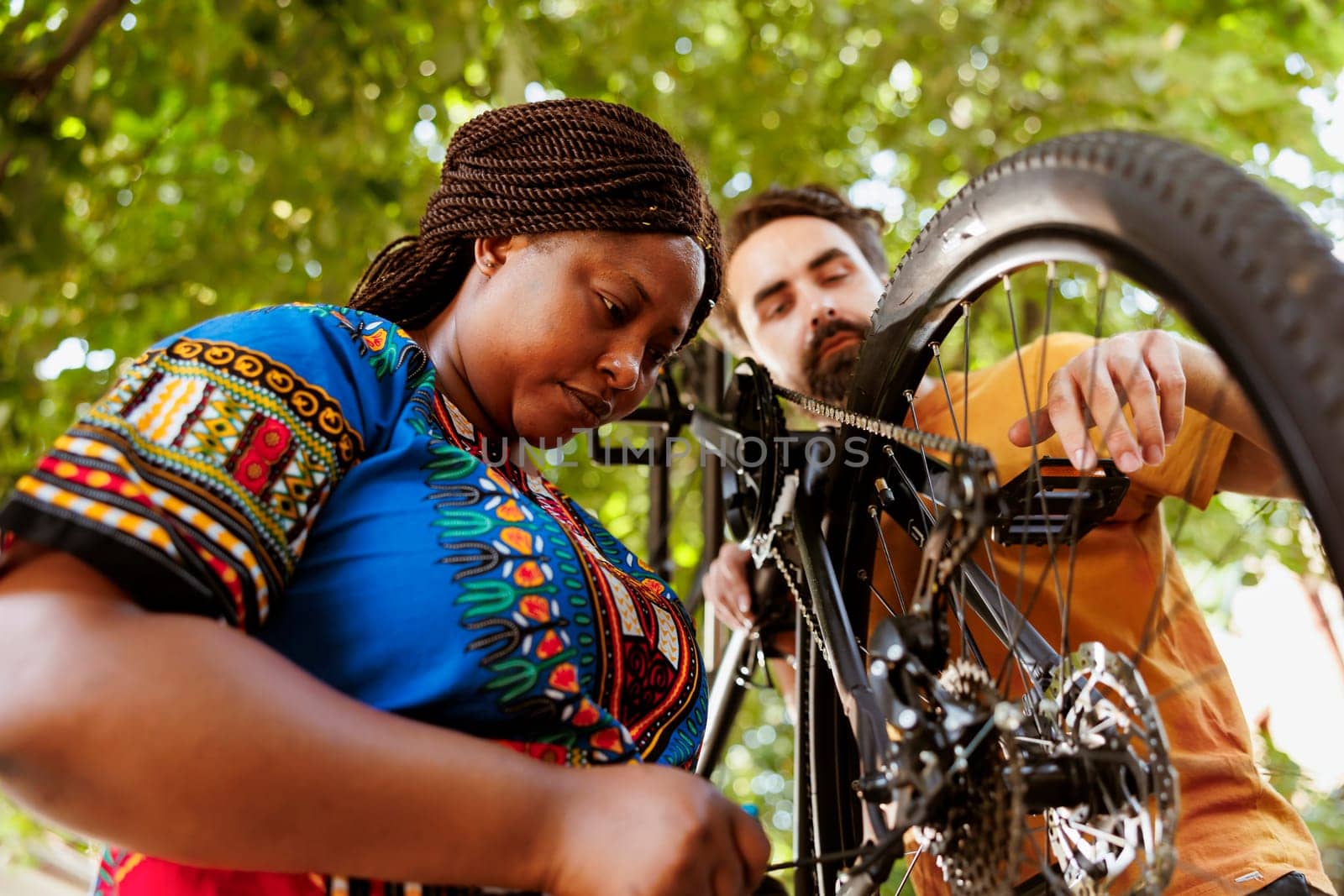 Sporty and eager black woman in yard repairing bike derailleur for summer recreational cycling. Healthy committed african american female cyclist assisted by caucasian man in repairing of bicycle components.