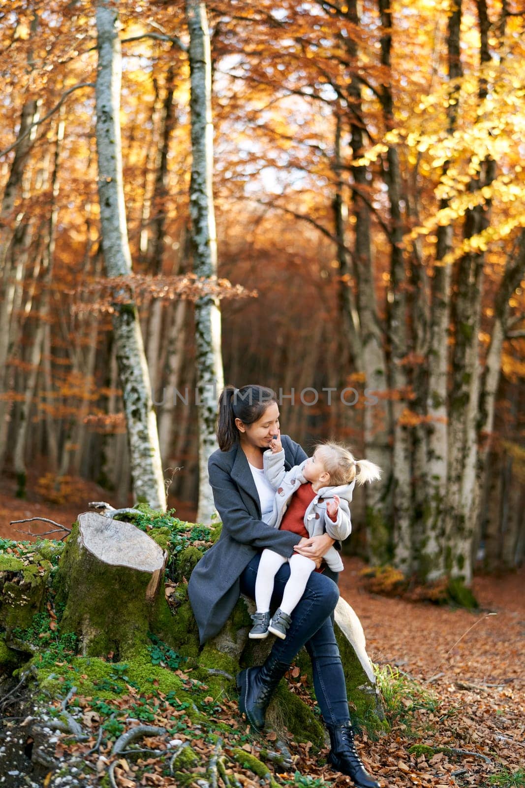 Little girl touches her mother cheek while sitting on her knees on a stump in the autumn forest. High quality photo