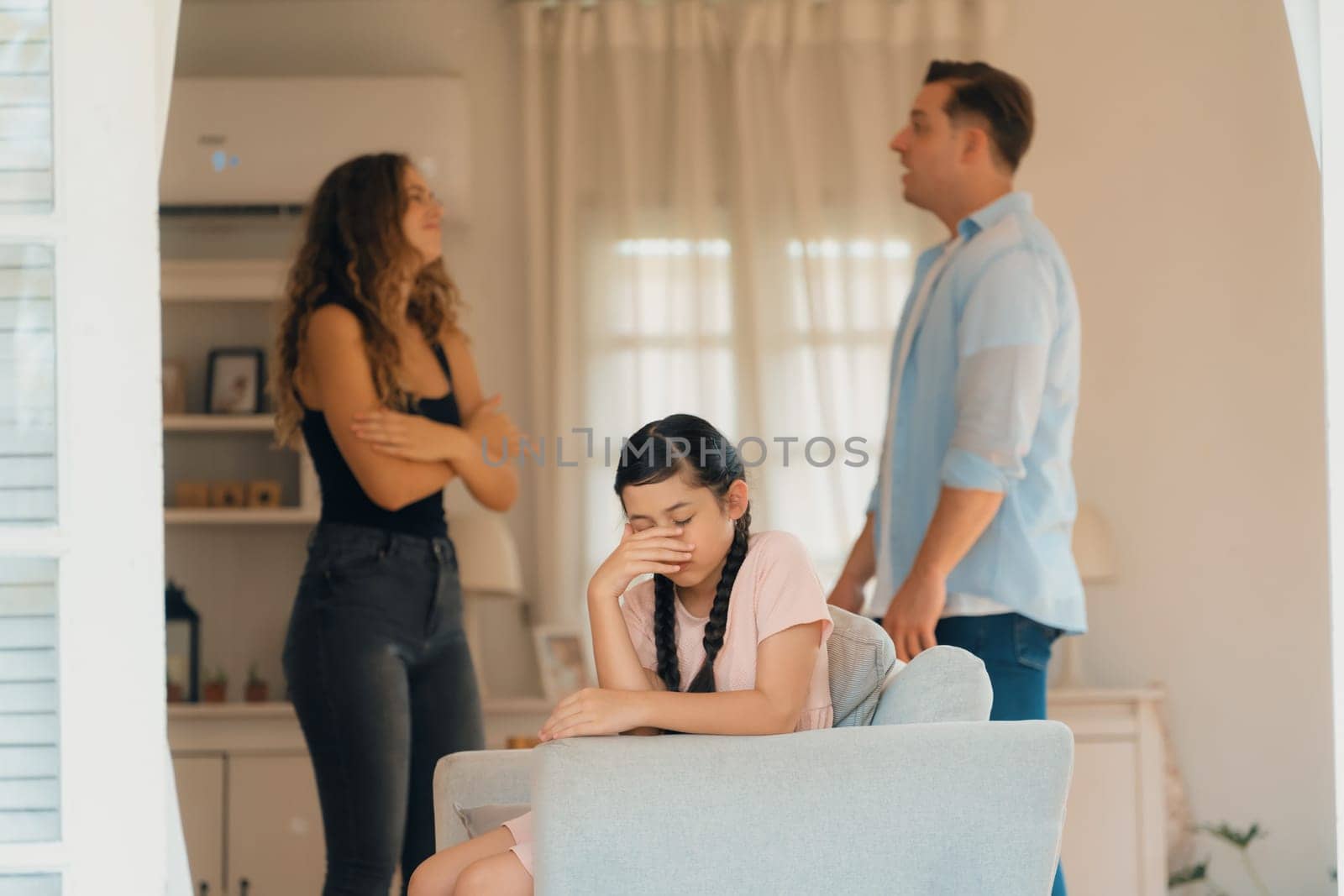 Annoyed and unhappy young girl sitting on sofa trapped in middle of tension by her parent argument in living room. Unhealthy domestic lifestyle and traumatic childhood develop to depression.Synchronos