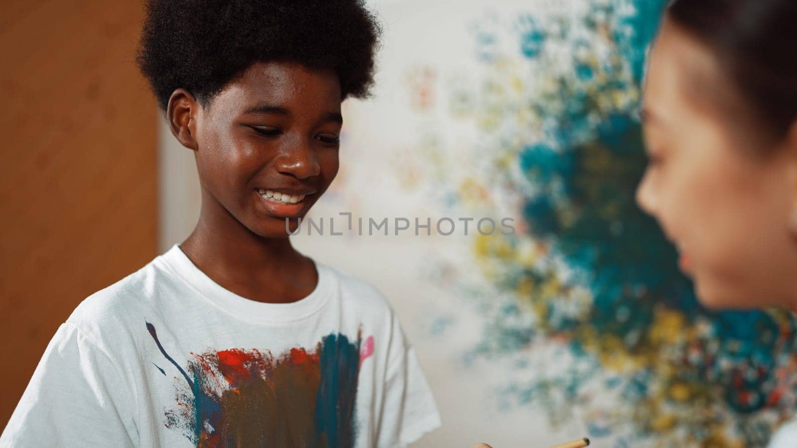 Smiling diverse children using paint brush painted color on each other white shirt shirt at colorful stained wall in art lesson. Represent exchanging experience, learning each other. Edification.