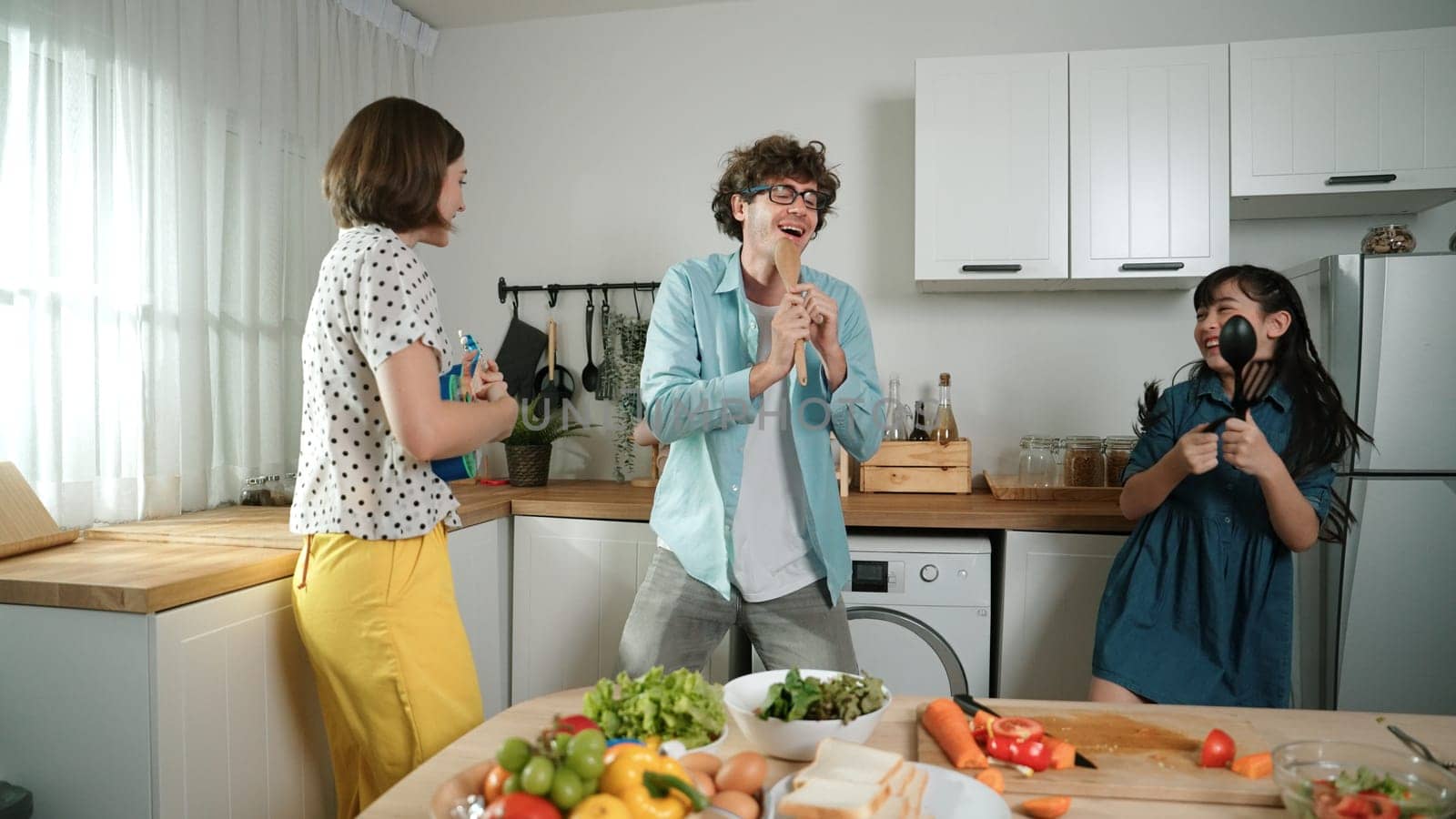 Caucasian skilled father, mother and asian daughter making breakfast while dancing together. Skilled mom playing ukulele while preparing vegetable at modern kitchen. Healthy food concept. Pedagogy.