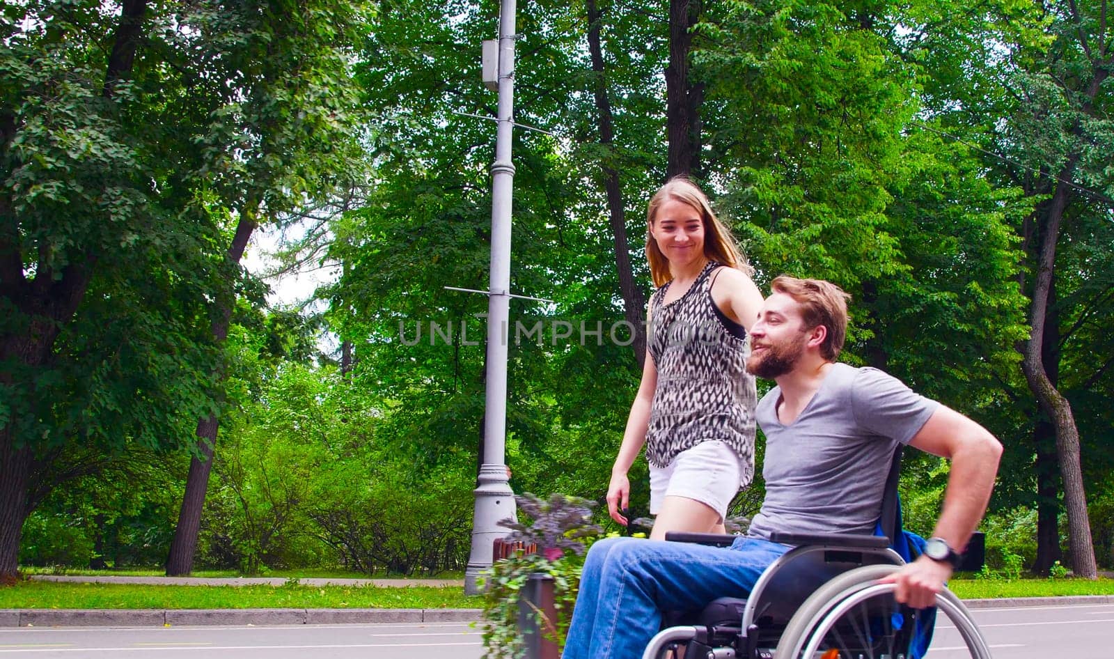 Happy young disable man in wheelchair walking in the park with his wife. Side view.