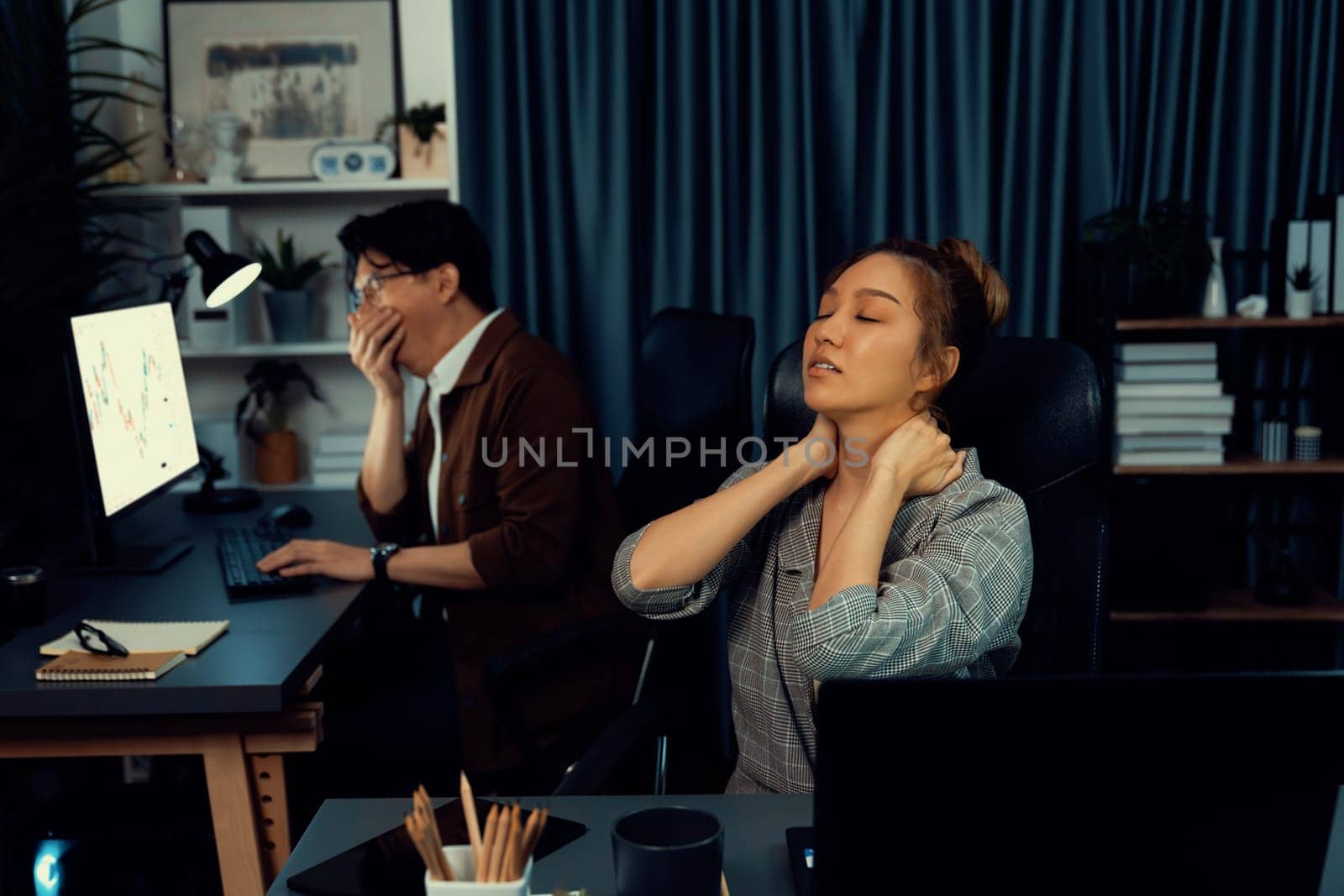 Businesswoman working on desk with stretching arm up and down manner with body health ache of strain overwork while coworker trading stock market on website on pc at late over night time. Infobahn.