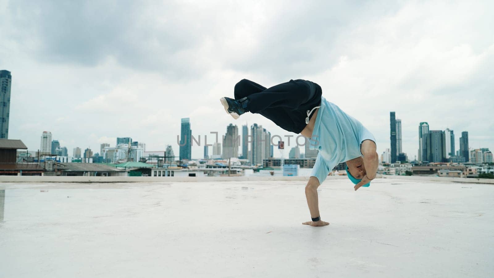 Caucasian B-boy dancer practicing street dancing at rooftop with city or urban. Motion shot of young man performing street dance by doing freeze pose or doing hand stand. Outdoor sport 2024. Endeavor.