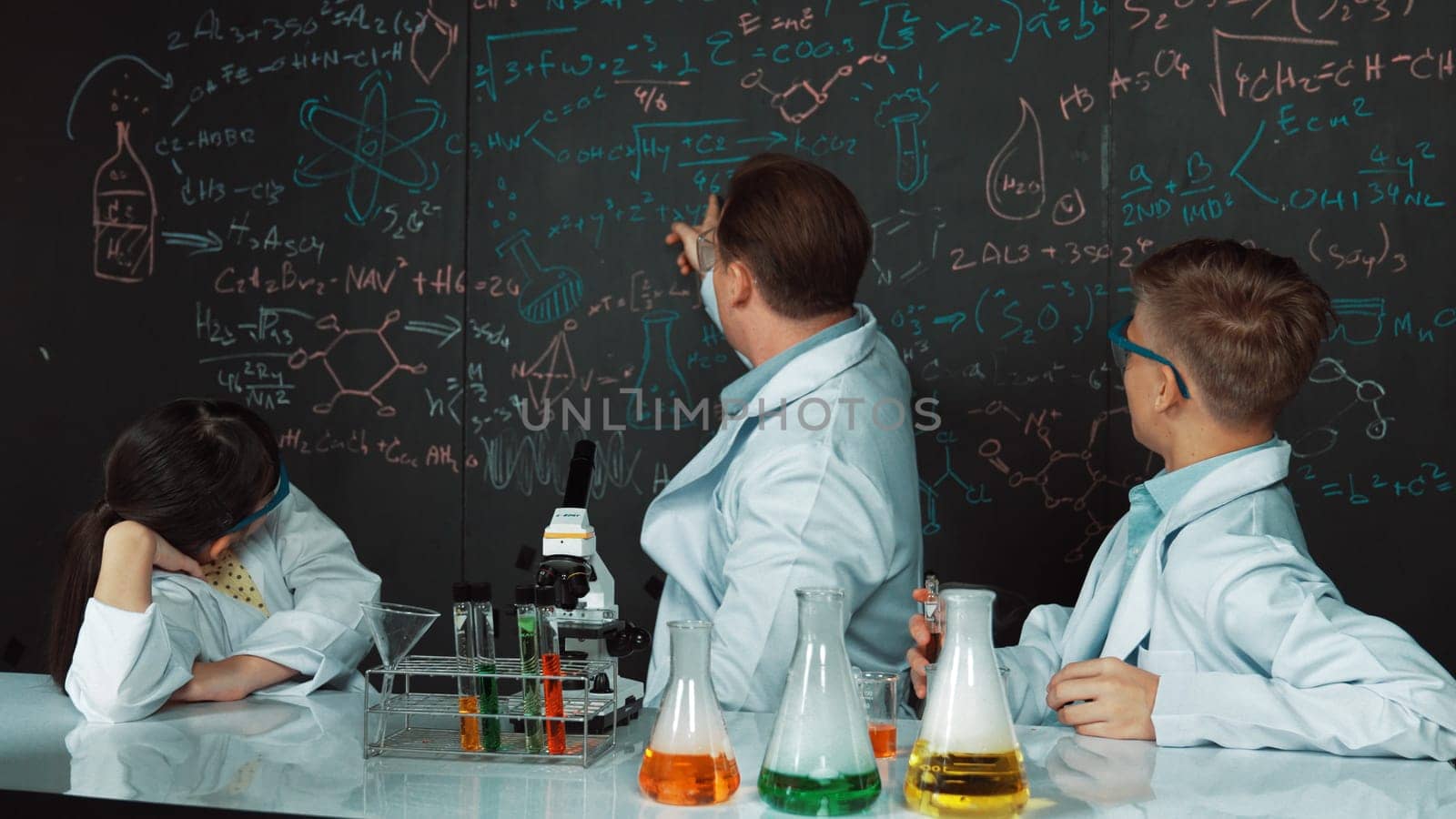 Creative teacher pointing chemistry at blackboard while talking to highschool boy at table with microscope and test tube with colored liquid. Young student wearing lab coat in STEM class. Edification.