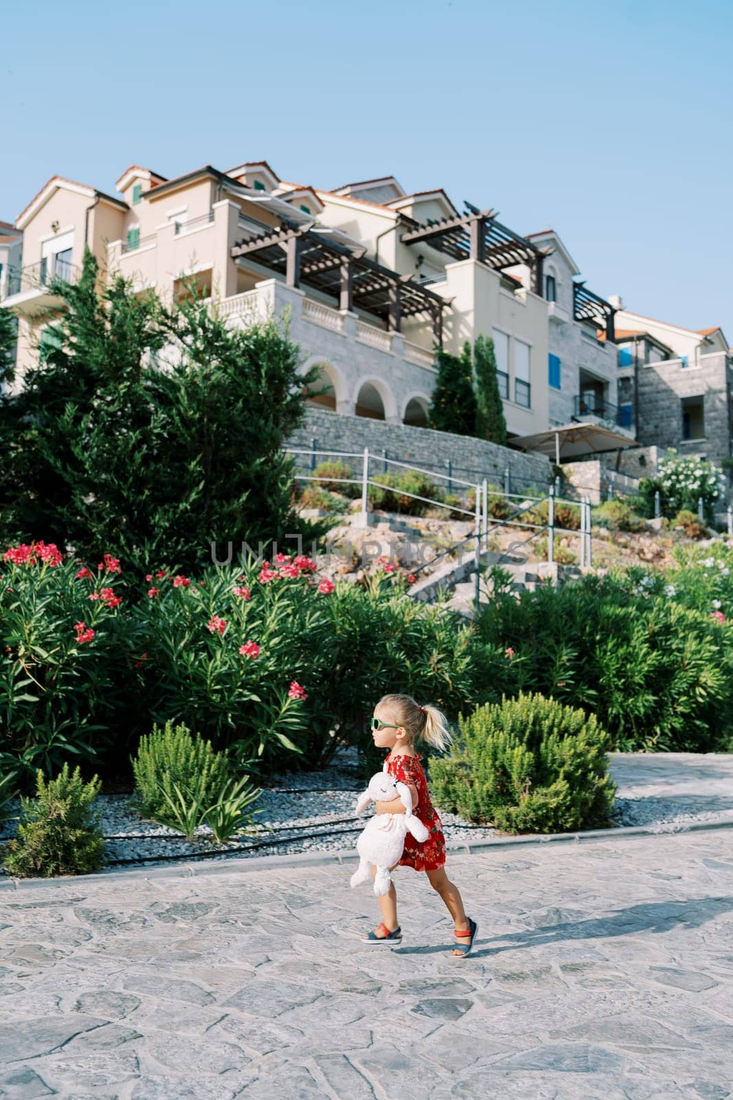 Little girl walks along a paved road in a flowering garden with stairs to a residential building with terraces. High quality photo