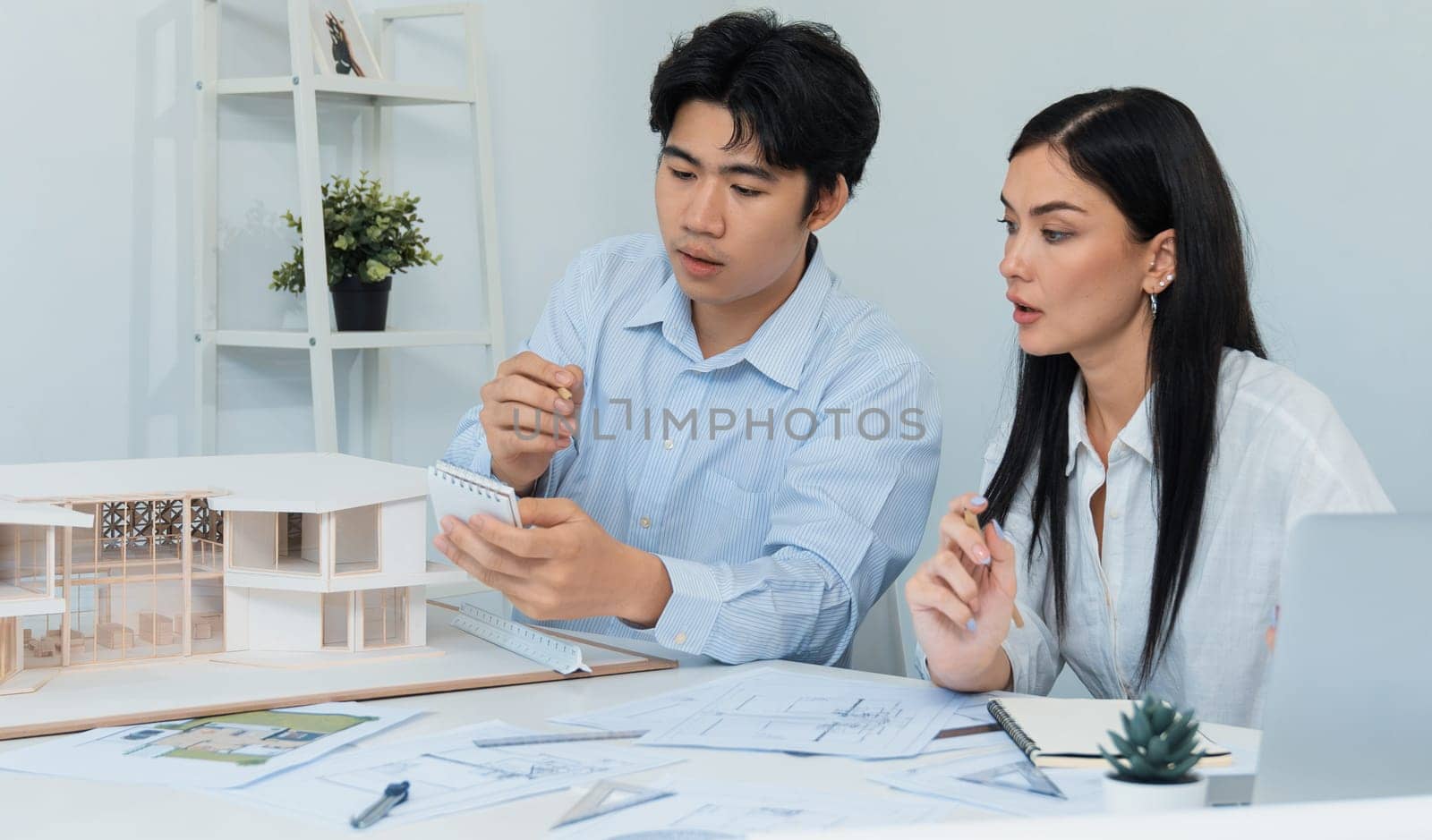 Professional male asian architect using ruler to measure house model length while young beautiful caucasian colleague using laptop to analyzed data on meeting table with house model. Immaculate.