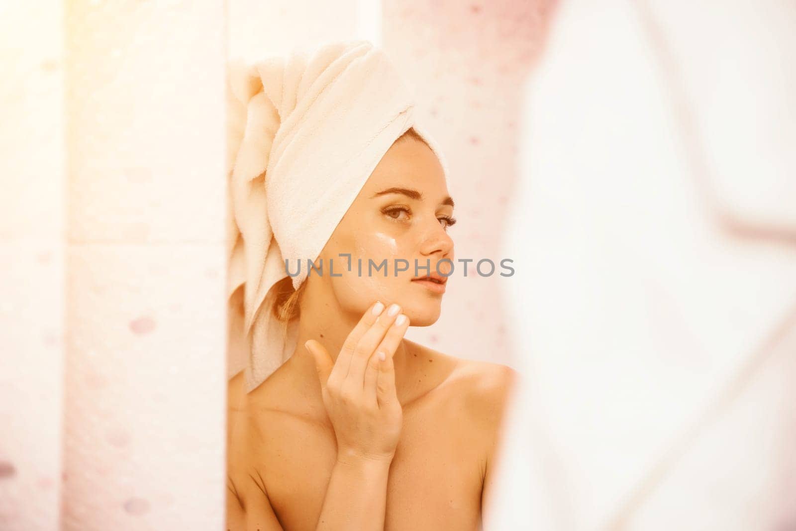 Young beautiful woman using face cream moisturizing lotion after bath. Pretty attractive girl in a towel on her head stands in front of a mirror in a home bathroom. Daily hygiene and skin care by Matiunina