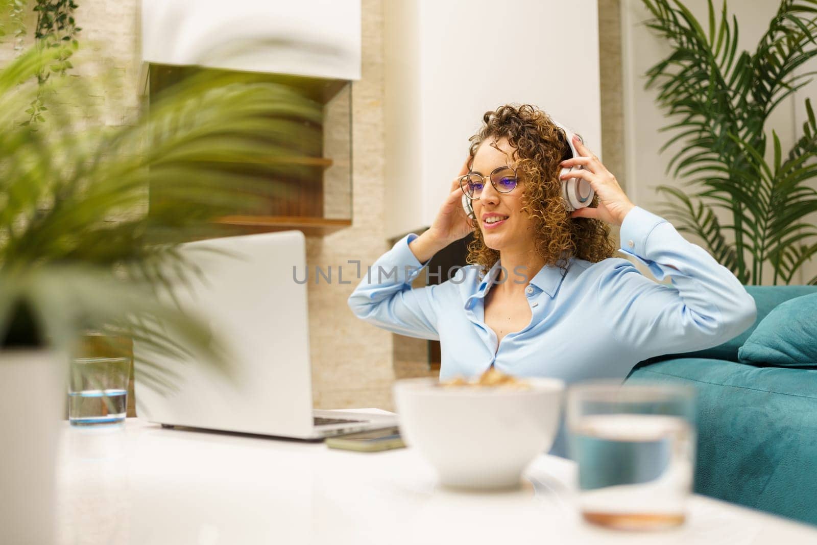 Positive young curly haired female freelancer, in smart casual shirt and eyeglasses listening to music over headphones while smiling and looking at screen of laptop in cozy living room