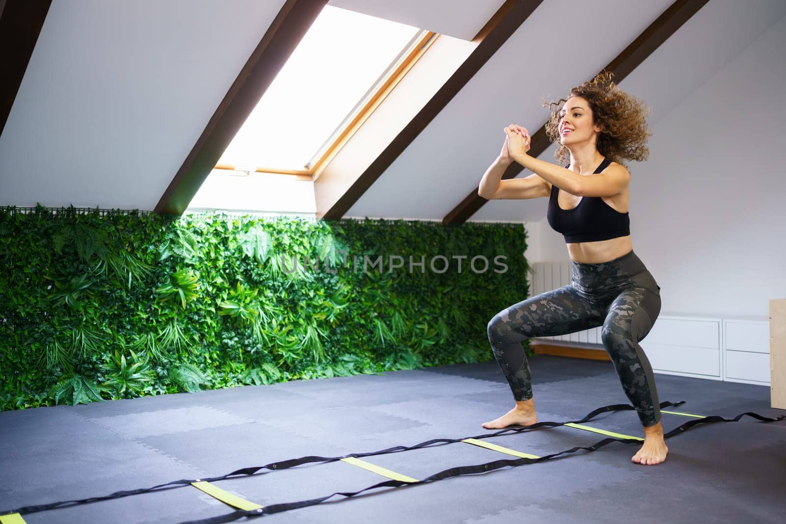 Full body of young curly haired female athlete in sportswear doing agility ladder squat jumps in gym with pitched roof