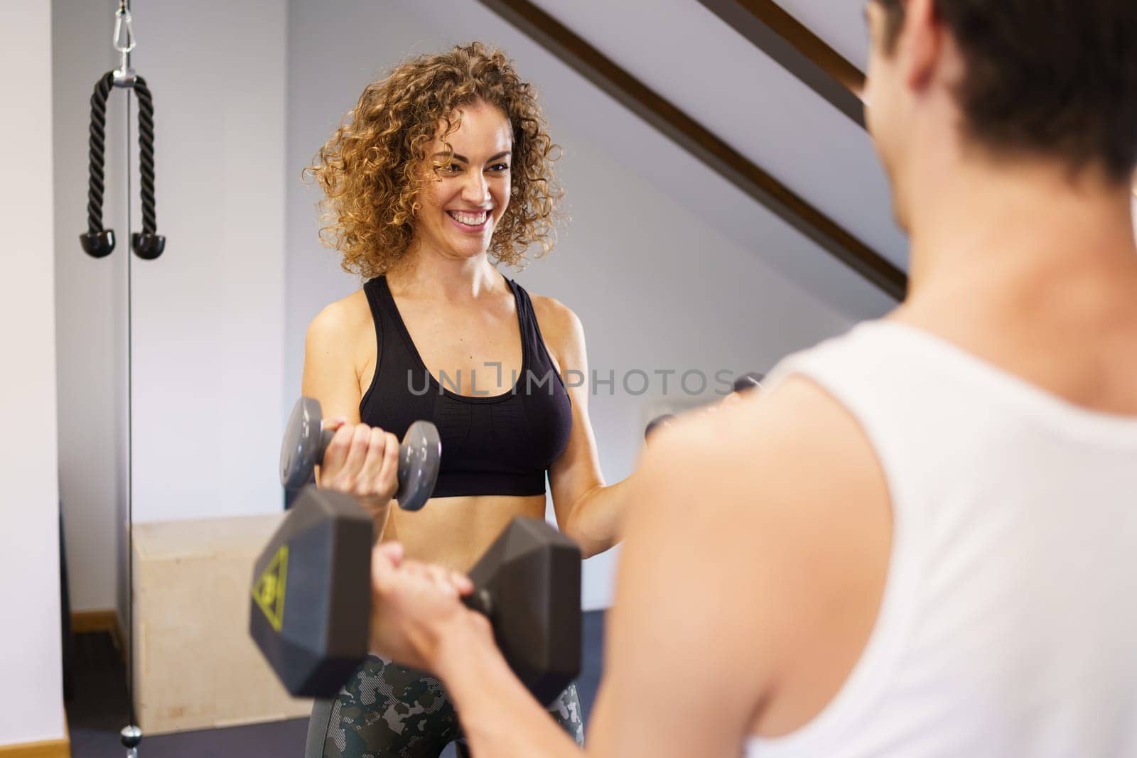 Positive fit woman in sports top lifting dumbbells with male friend while pumping muscles during fitness training in gym
