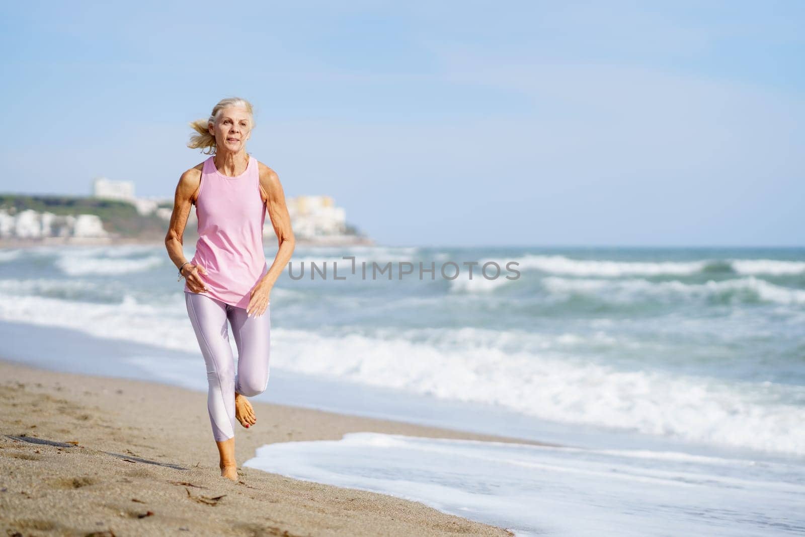 Older female doing sport to keep fit. Mature woman running along the shore of the beach. Concept of healthy living in the elderly. Senior woman in fitness clothing running along beach