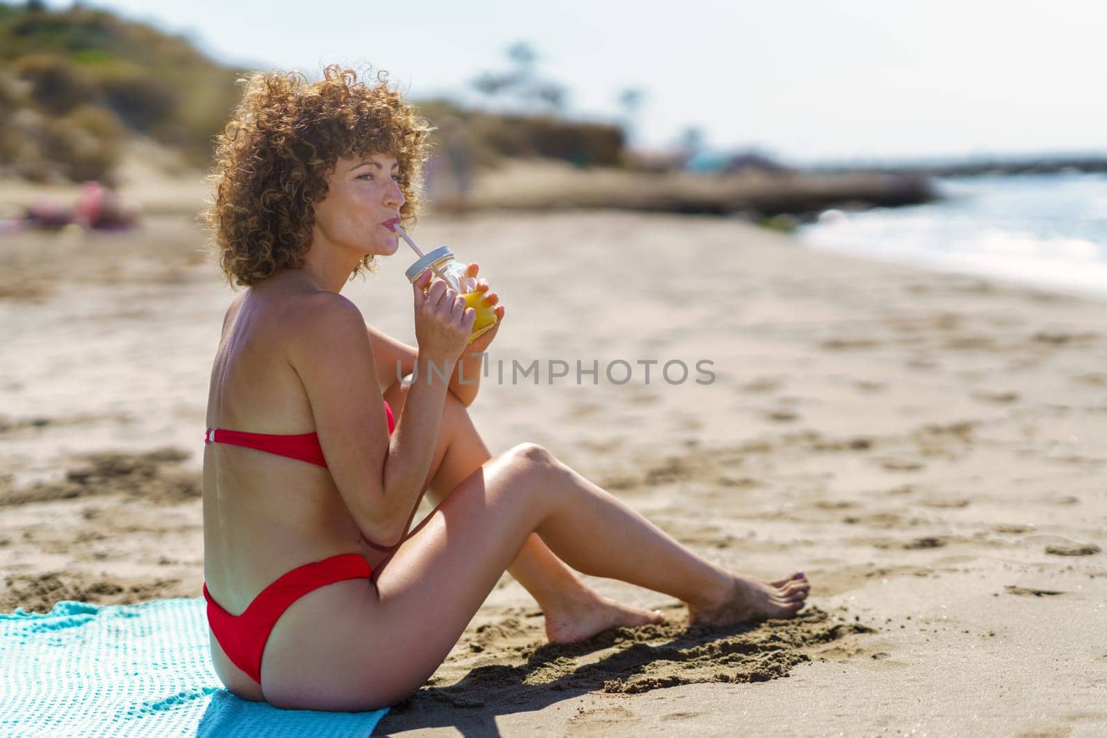 Side view of dreamy curly woman in bikini sipping cool orange juice with straw while sitting on coast of ocean