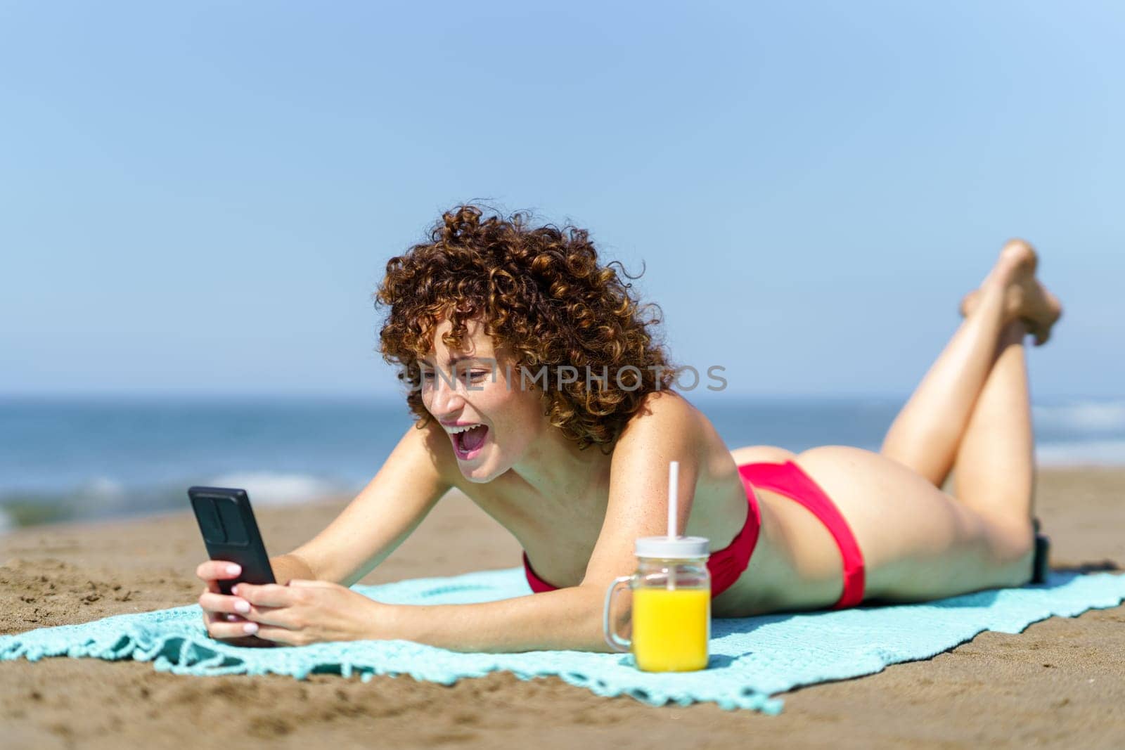 Laughing young curly haired lady in bikini having cup of cool drink while surfing smartphone and resting on sandy beach