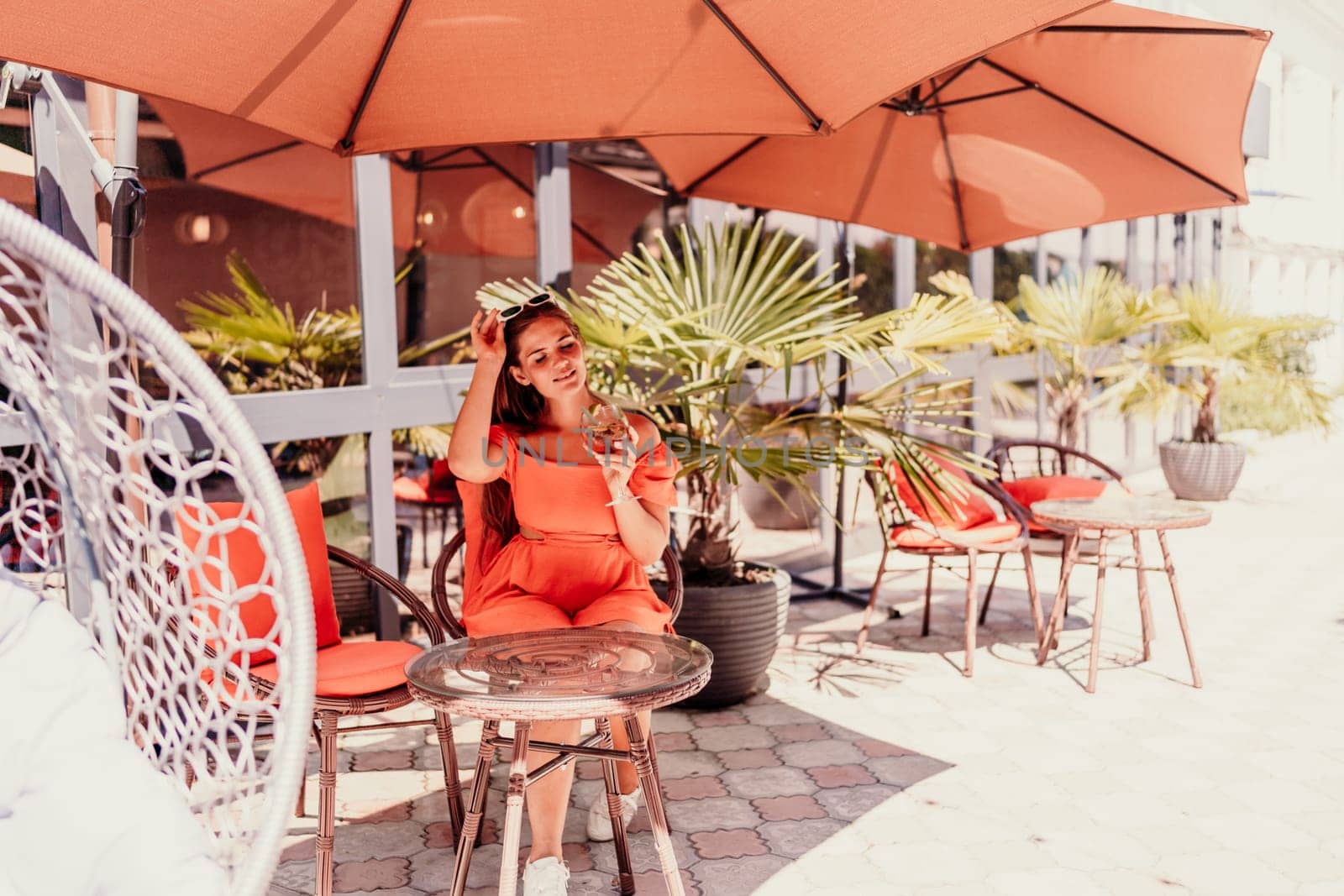 A woman in an orange dress sits at a table with a drink in a glass. The scene is set in a restaurant or cafe, with a potted plant nearby. The woman is enjoying her time, and the atmosphere is relaxed. by Matiunina