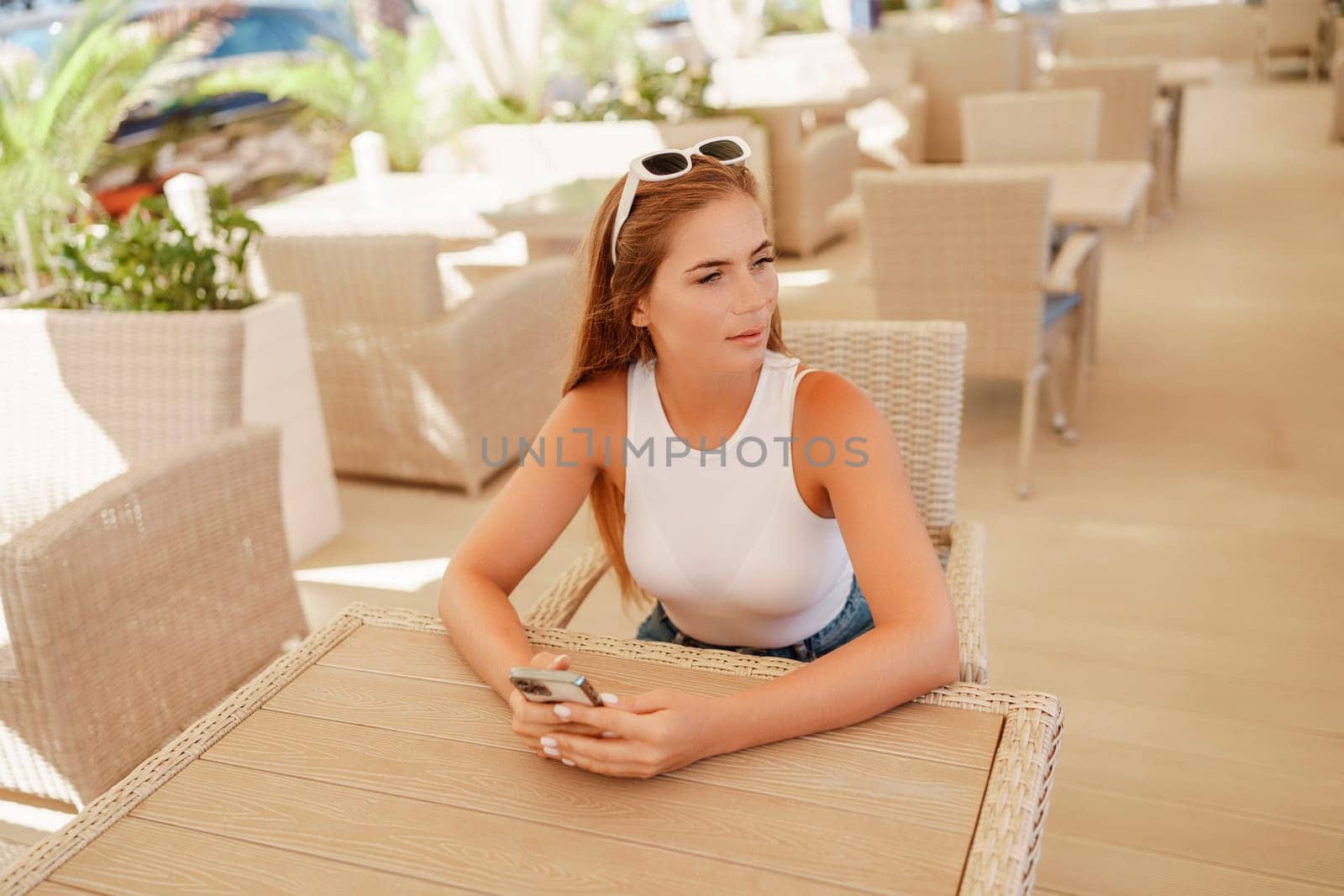 Woman sits in outdoor cafe at wooden table, holds smartphone. Bright sunny day provides natural lighting. Cafe offers relaxed atmosphere for customers to enjoy refreshments. by Matiunina