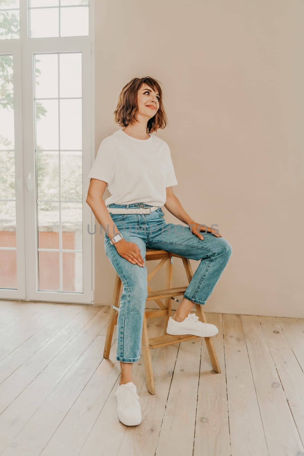 Portrait of brunette white t-shirt. A woman sits on a chair and looks at the camera, business portrait on a beige background, dressed in a white t-shirt and jeans. by Matiunina