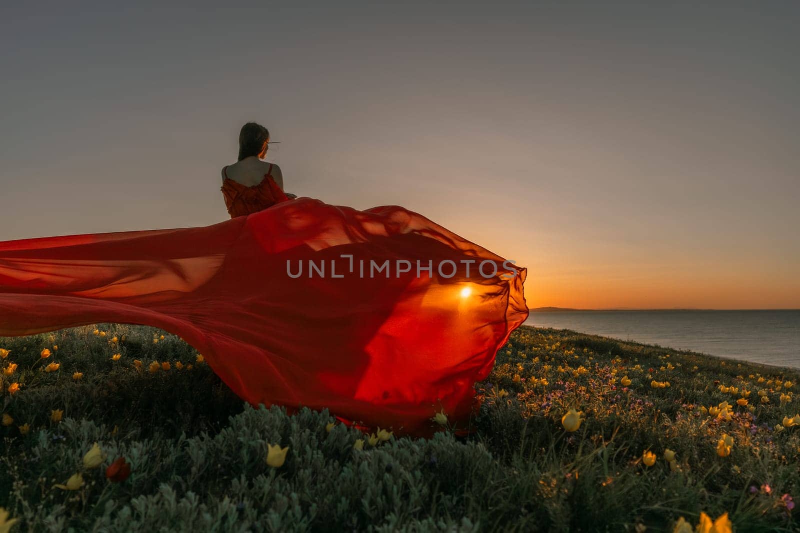 A woman in a red dress is standing in a field with the sun setting behind her. She is reaching up with her arms outstretched, as if she is trying to catch the sun. The scene is serene and peaceful. by Matiunina