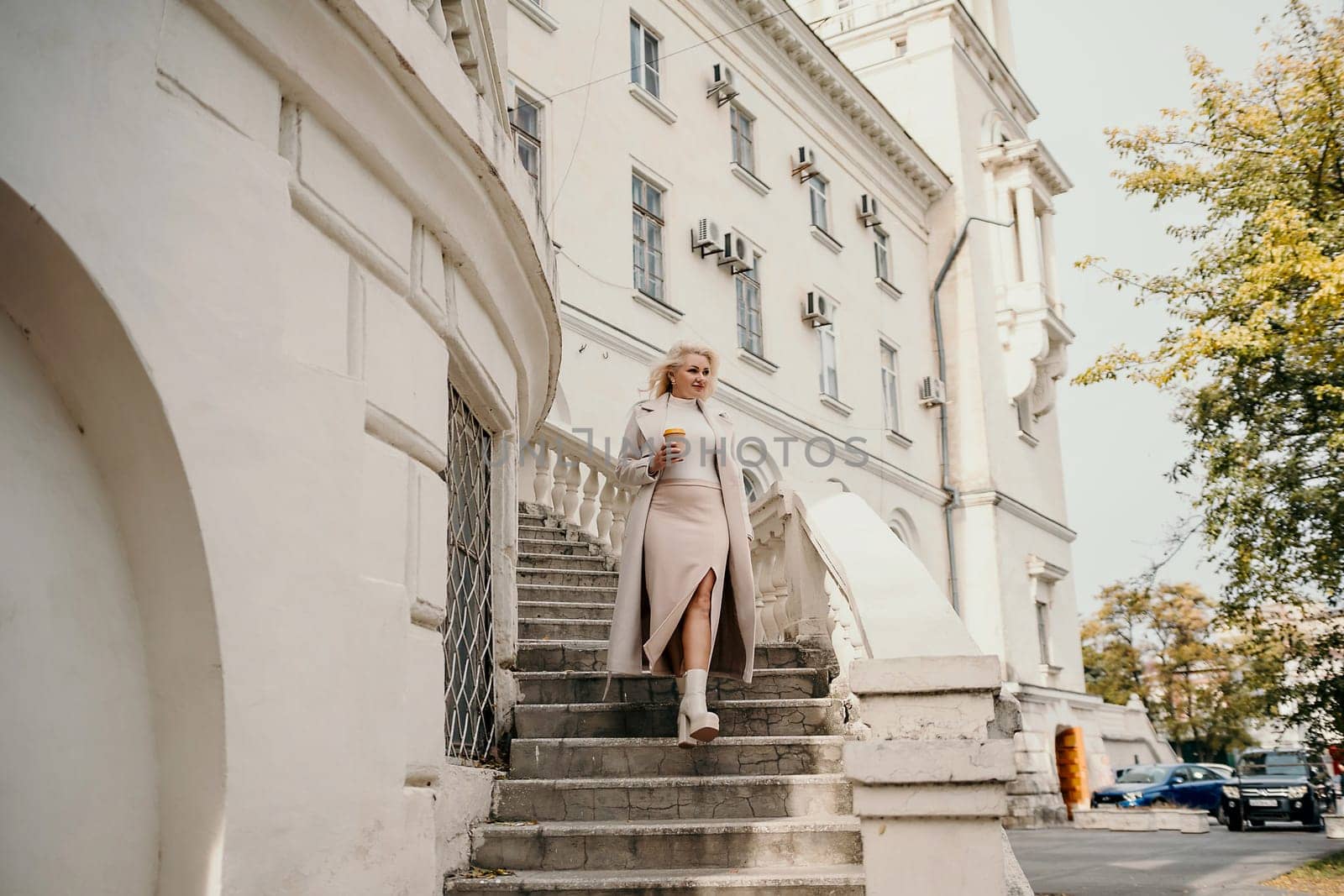A woman in a white dress is walking up a set of stairs. She is holding a cup in her hand. The scene is set in front of a large white building with a balcony