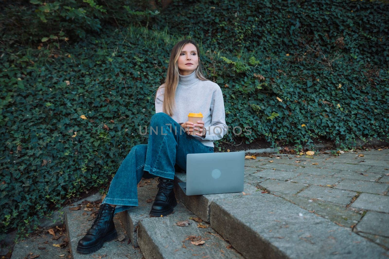 A woman sits on the ground with a cup in her hand. She is wearing a gray sweater and blue jeans. by Matiunina