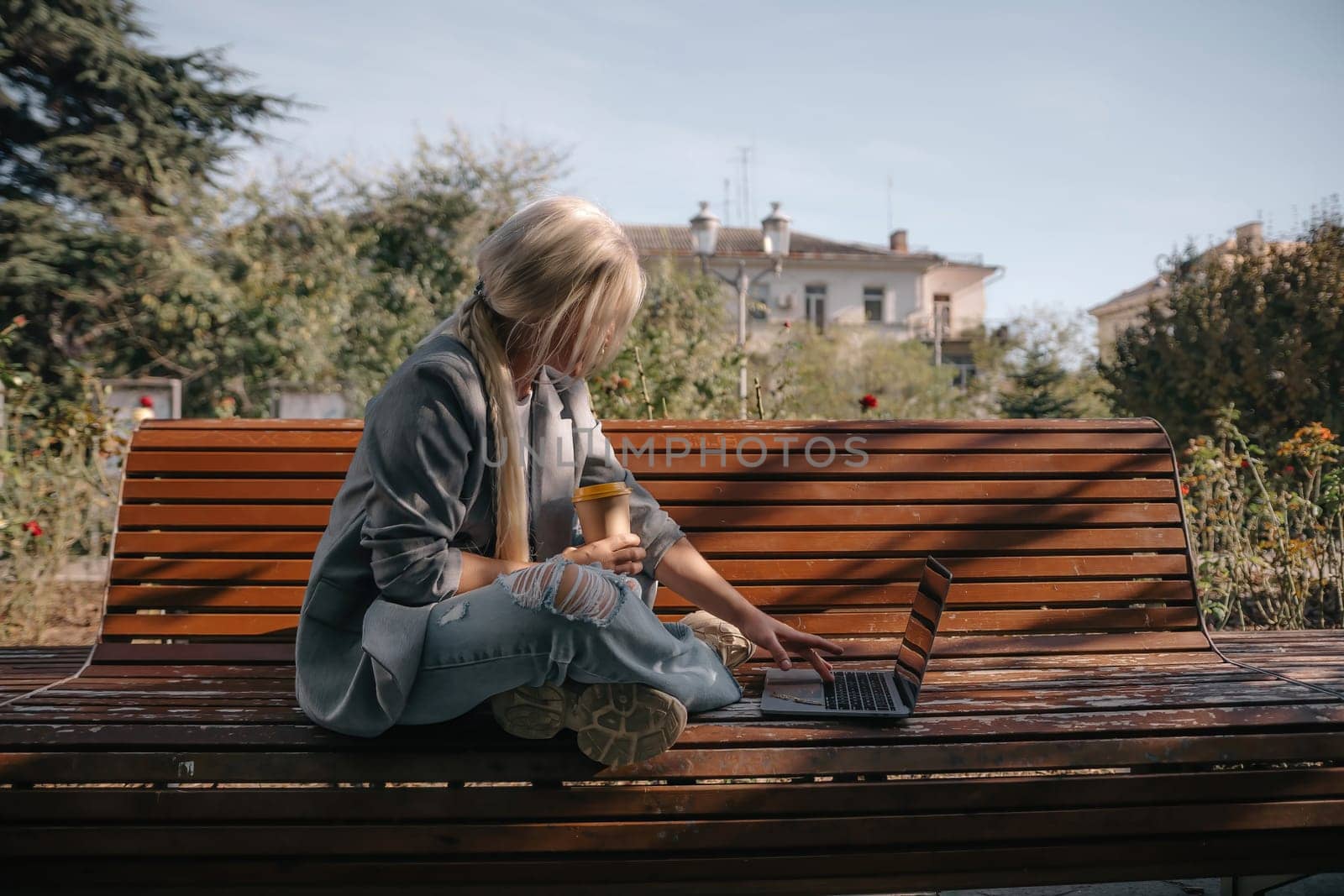 A woman sits on a bench with a laptop open in front of her. She has a cup of coffee in her hand and her jeans are ripped. The scene suggests a relaxed and casual atmosphere