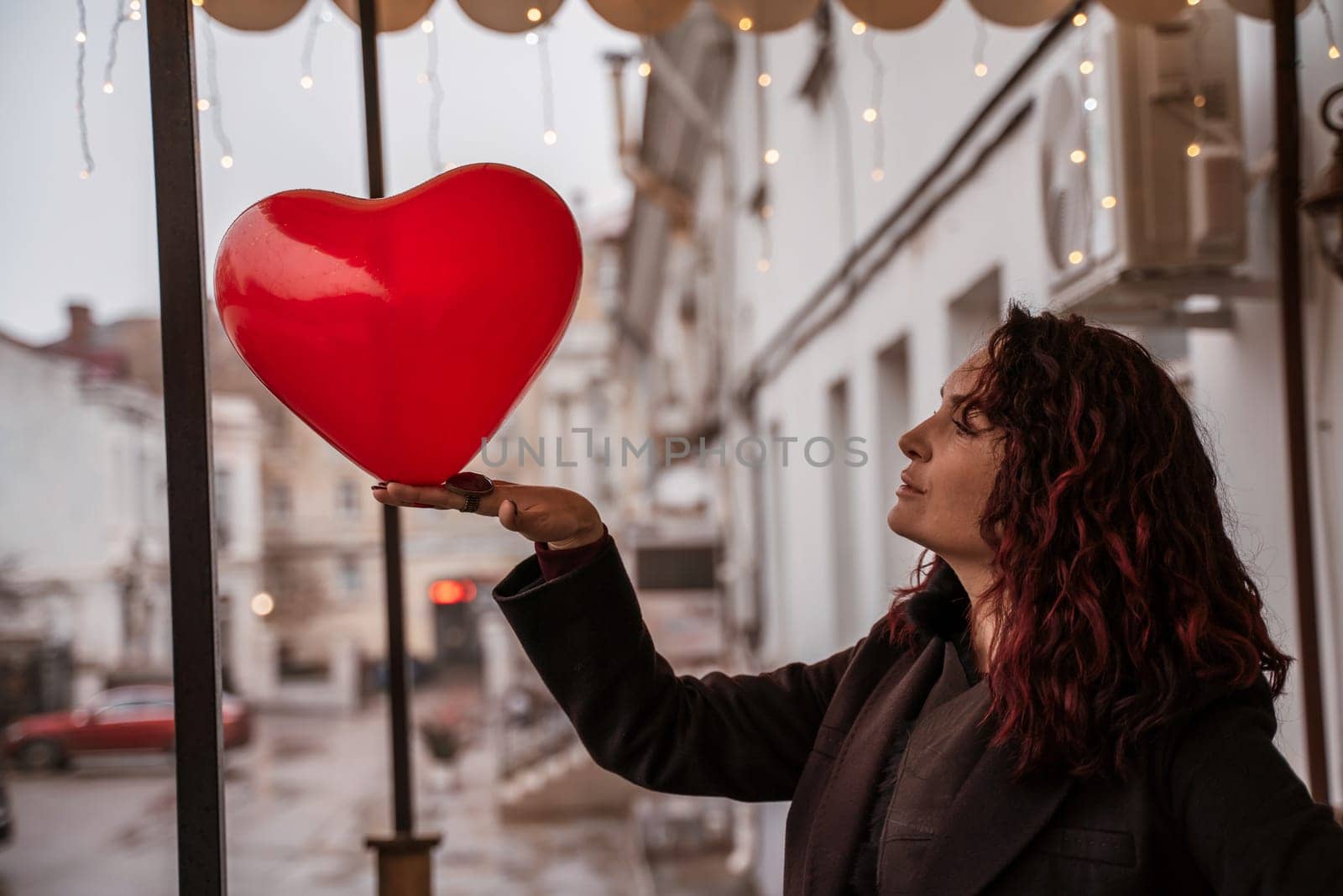 A woman holding a red heart balloon. The woman is wearing a black coat and has red hair. The scene is set in a city street with cars and a building in the background