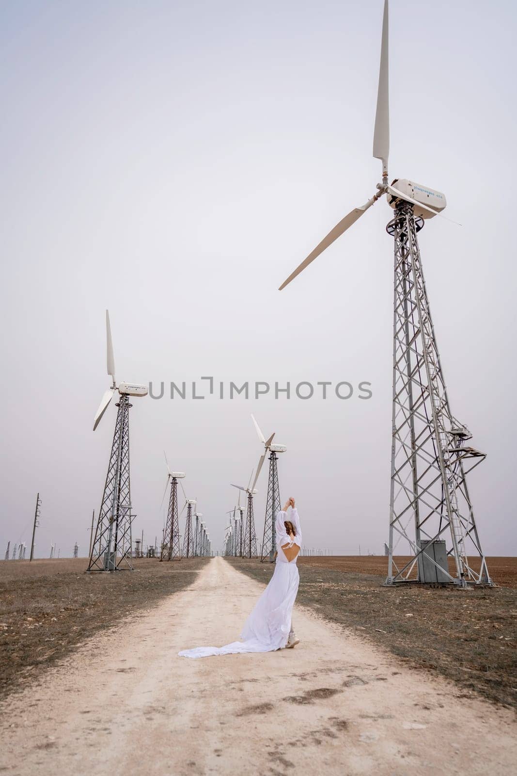 A woman in a white dress is walking down a dirt road in front of a row of wind turbines