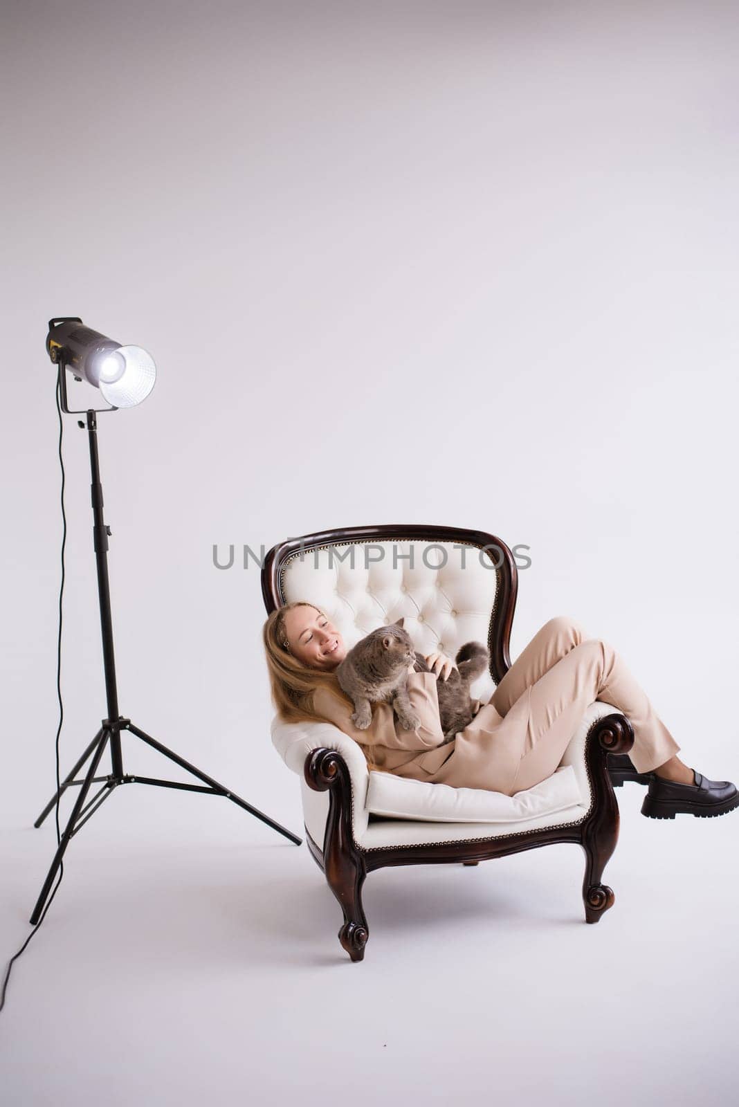 Female blogger, blonde on chair with domestic cat scottish straight in white photo studio. Dressed in a formal beige trouser suit. Vertical
