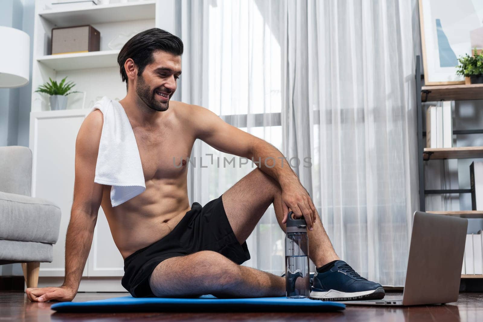 Athletic and sporty man resting on fitness mat during online body workout exercise session for fit physique and healthy sport lifestyle at home. Gaiety home exercise workout training concept.