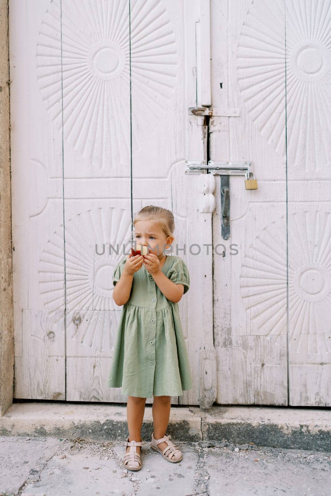 Little girl gnaws a big apple while standing near a wooden antique carved door by Nadtochiy