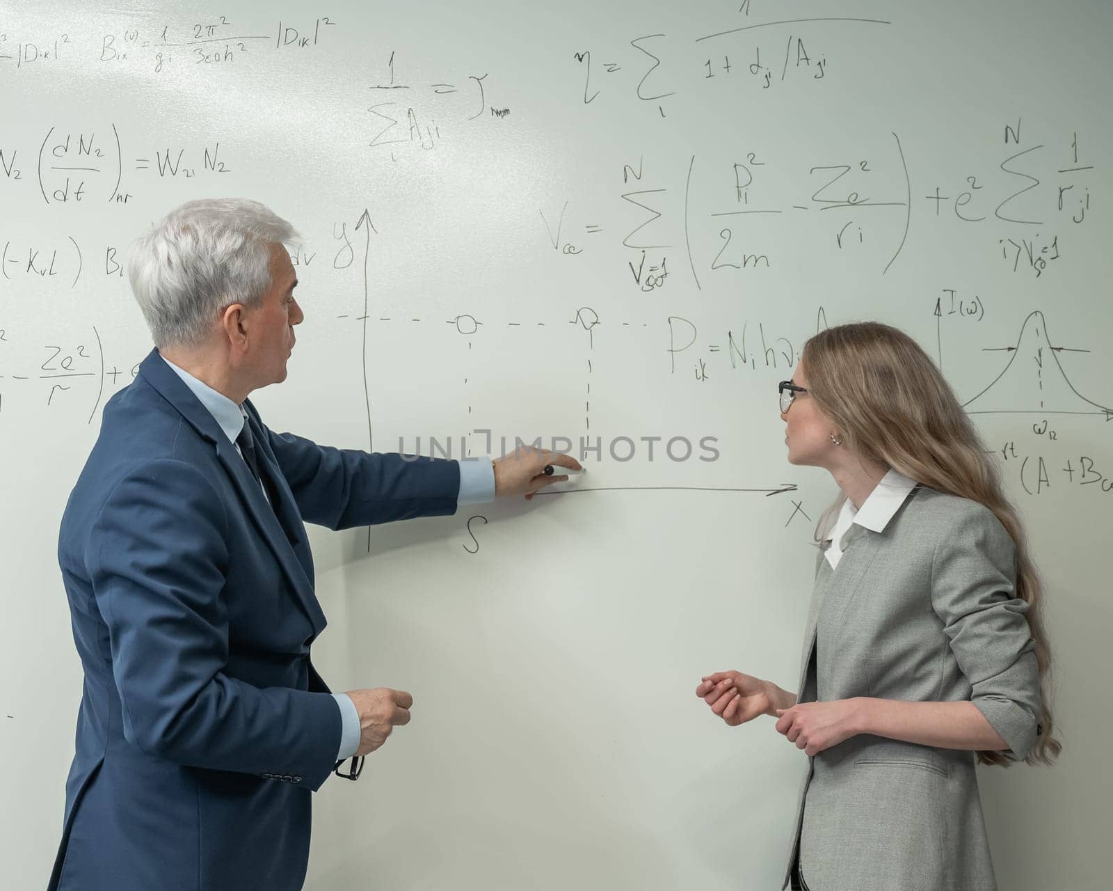 An elderly professor explains a subject to a student at a white board. by mrwed54