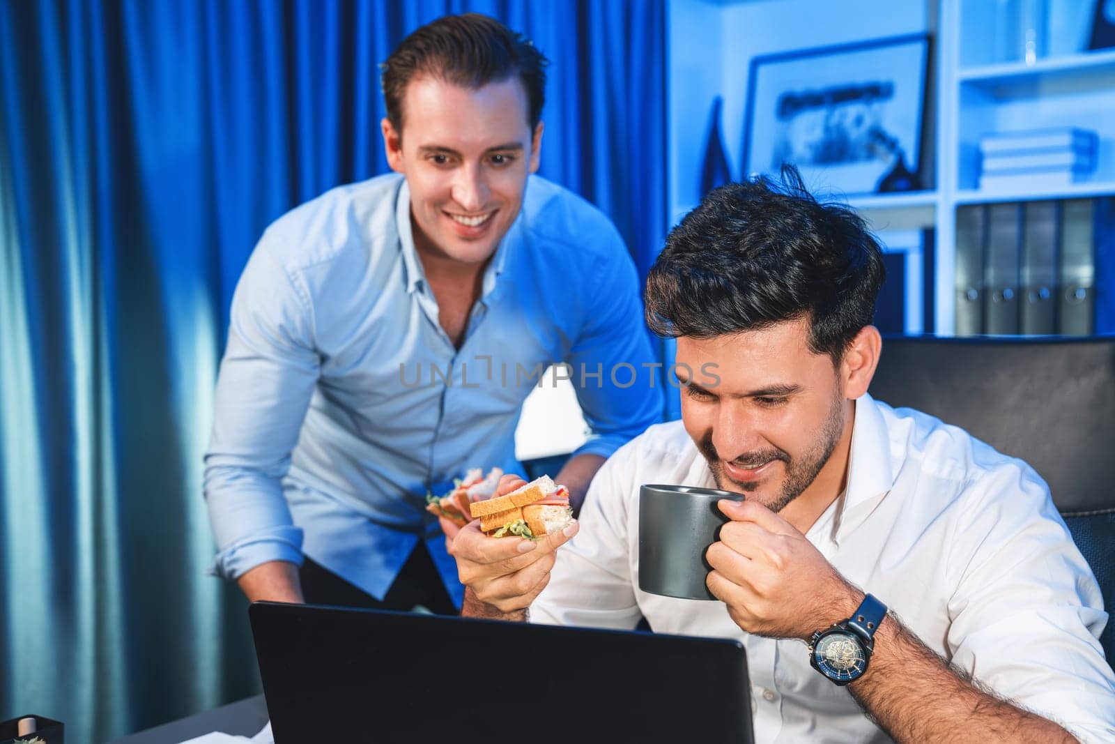 Business partners with happy smiling face sharing delicious sandwich dish with friend at night time. Hungry creative colleagues enjoy eating fast food at workplace with neon blue light. Sellable.